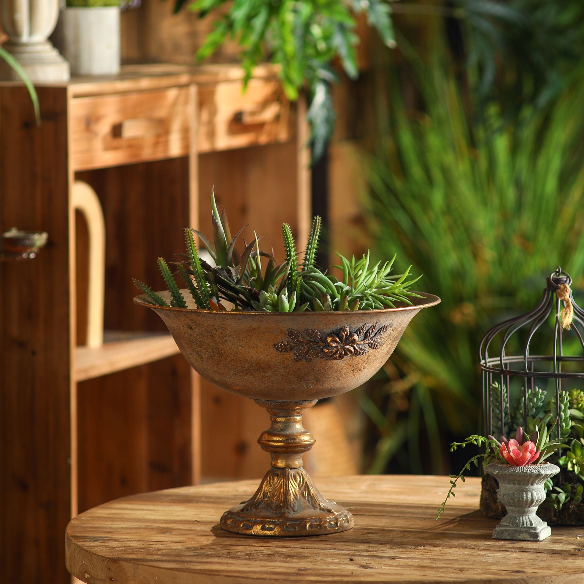Orlando compote bowl in antique gold with flower garland on a round table with statue behind