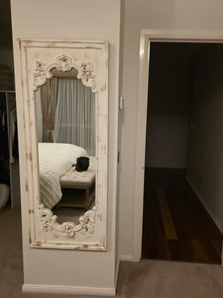 A tall, distressed white wooden floor mirror with ornate carved corners, leaning against a wall in a bedroom hallway next to an open doorway.