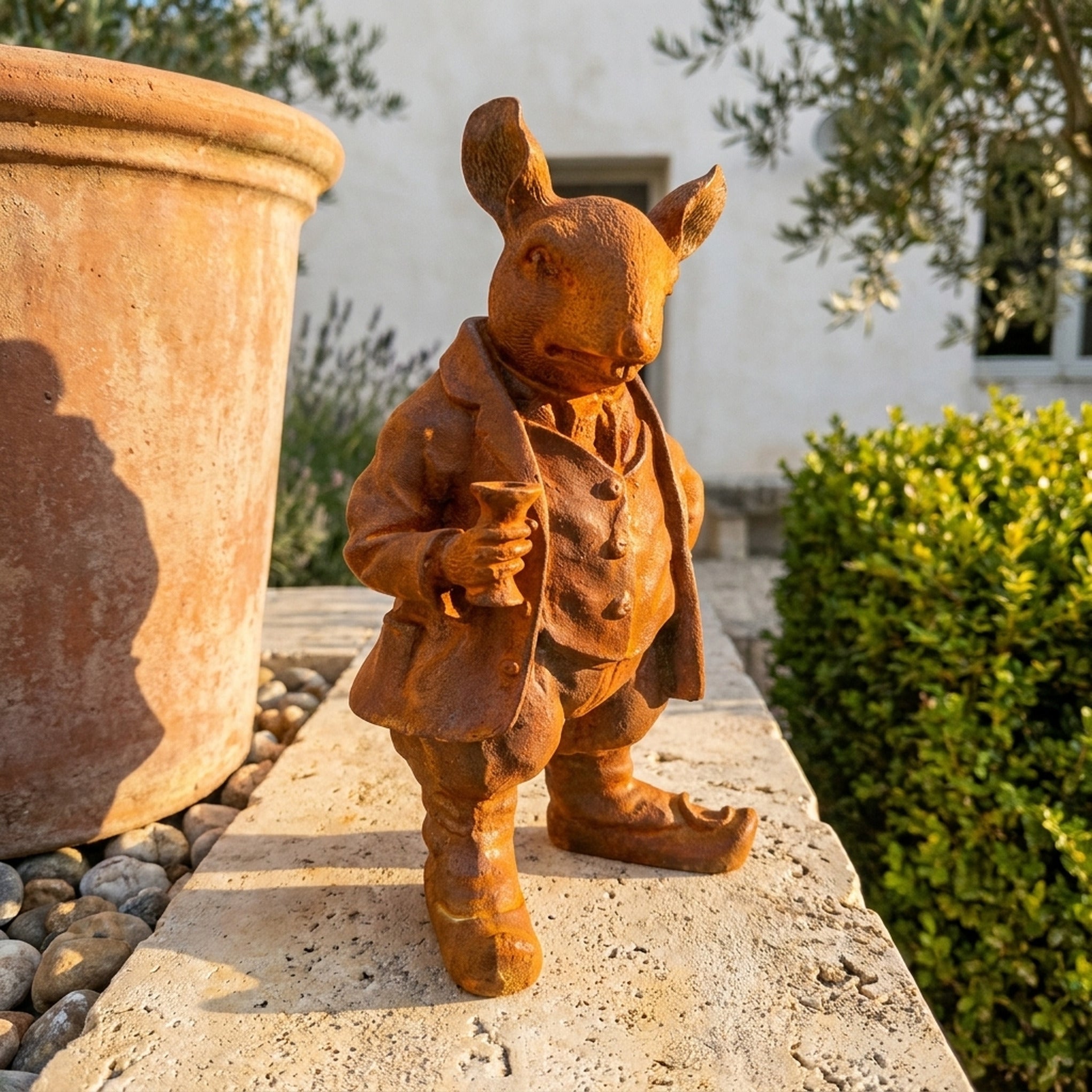 Monsieur Ratsworth rust cast iron rat statue in a frock coat raising a goblet, on a travertine ledge beside a terracotta pot