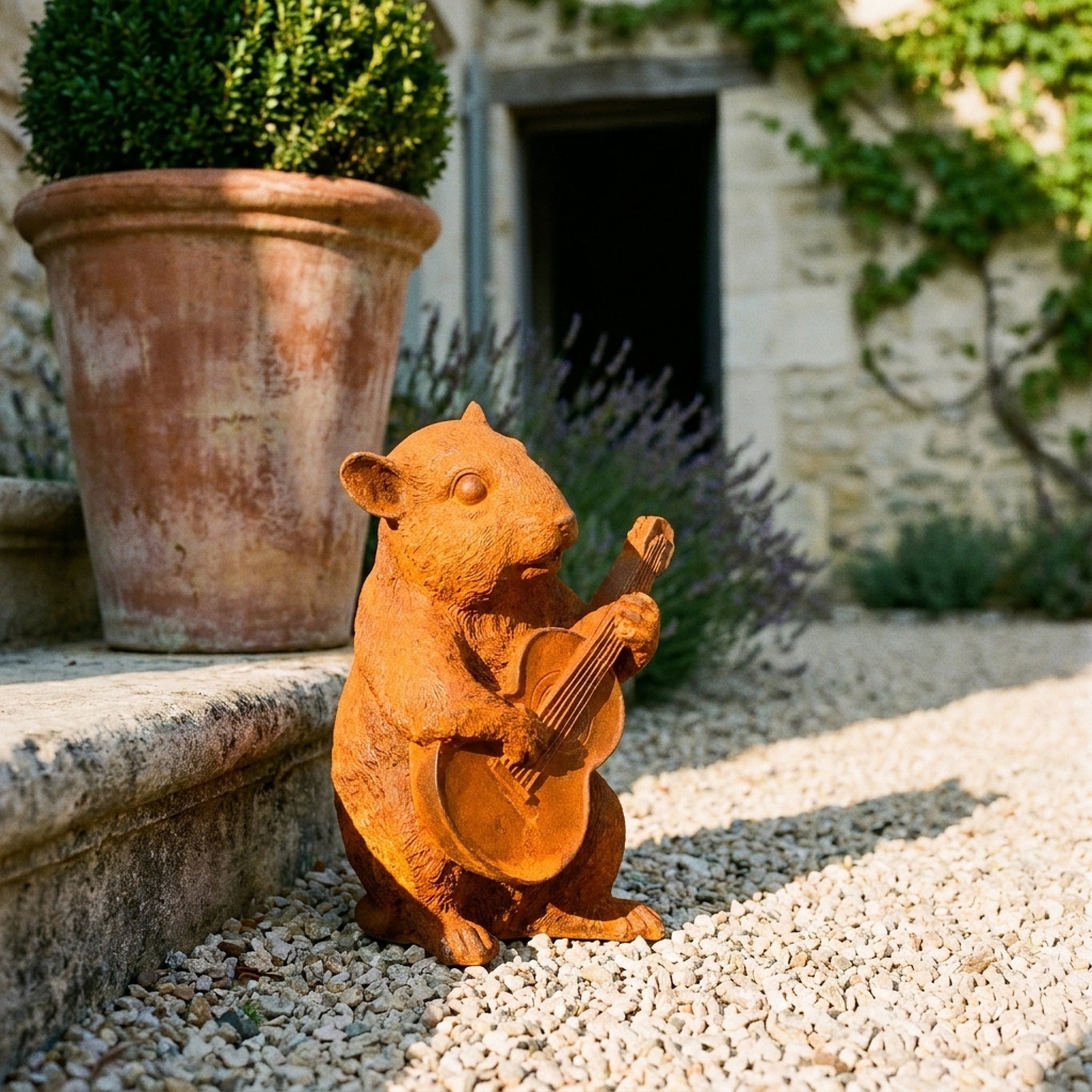 Bertie rust cast iron dormouse statue playing a guitar, on gravel beside a terracotta pot