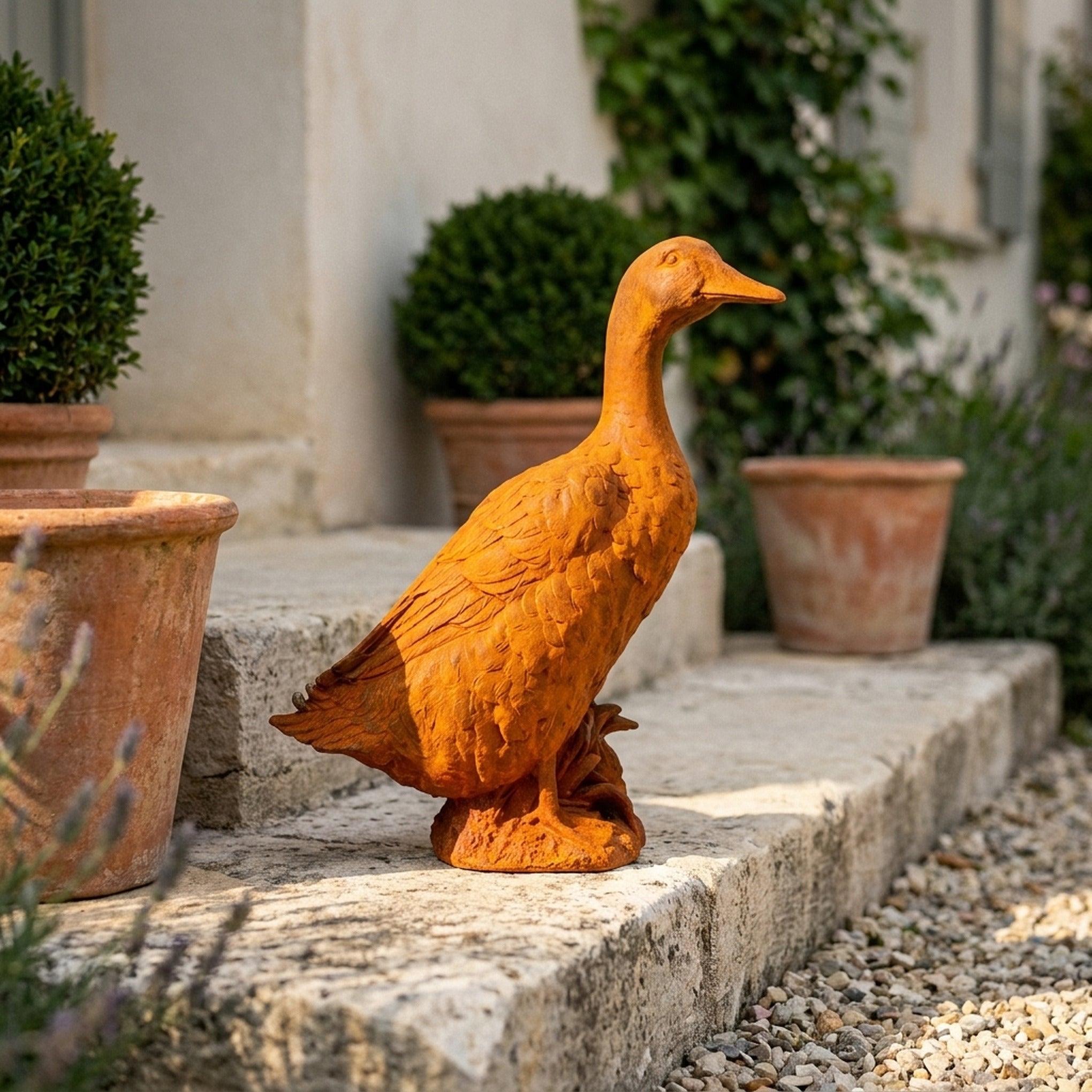 Goosey Gander rust cast iron duck statue on a rock base, standing on gravel beside a terracotta pot