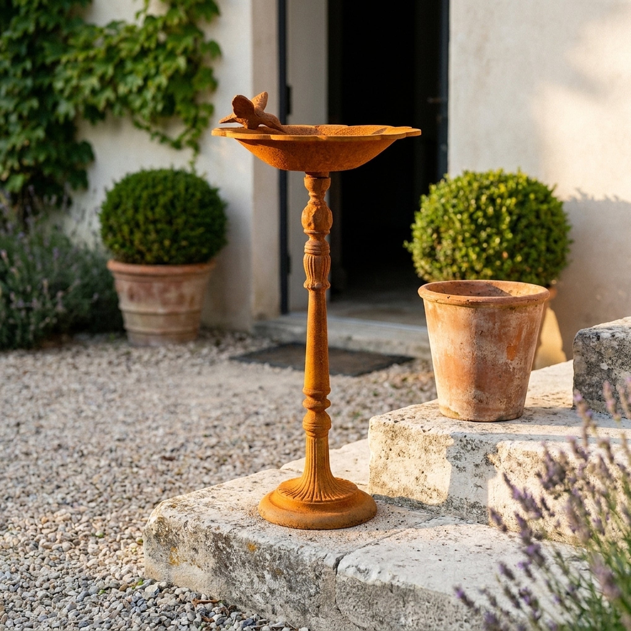 The Willowmere rust cast iron pedestal bird bath with a bird on the rim, standing on gravel in a courtyard