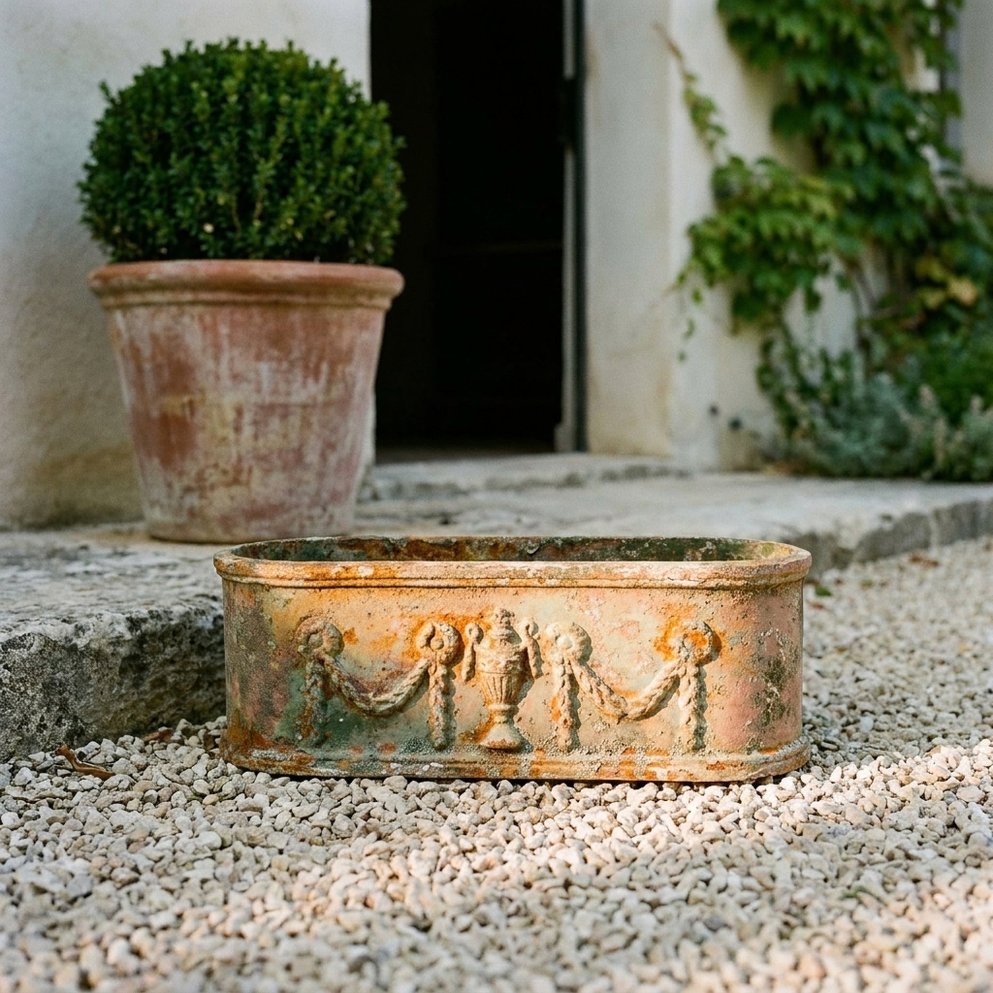 The Heritage Planter in weathered antique bronze cast iron, an oval trough with urn and swag relief, on gravel in a provencal courtyard