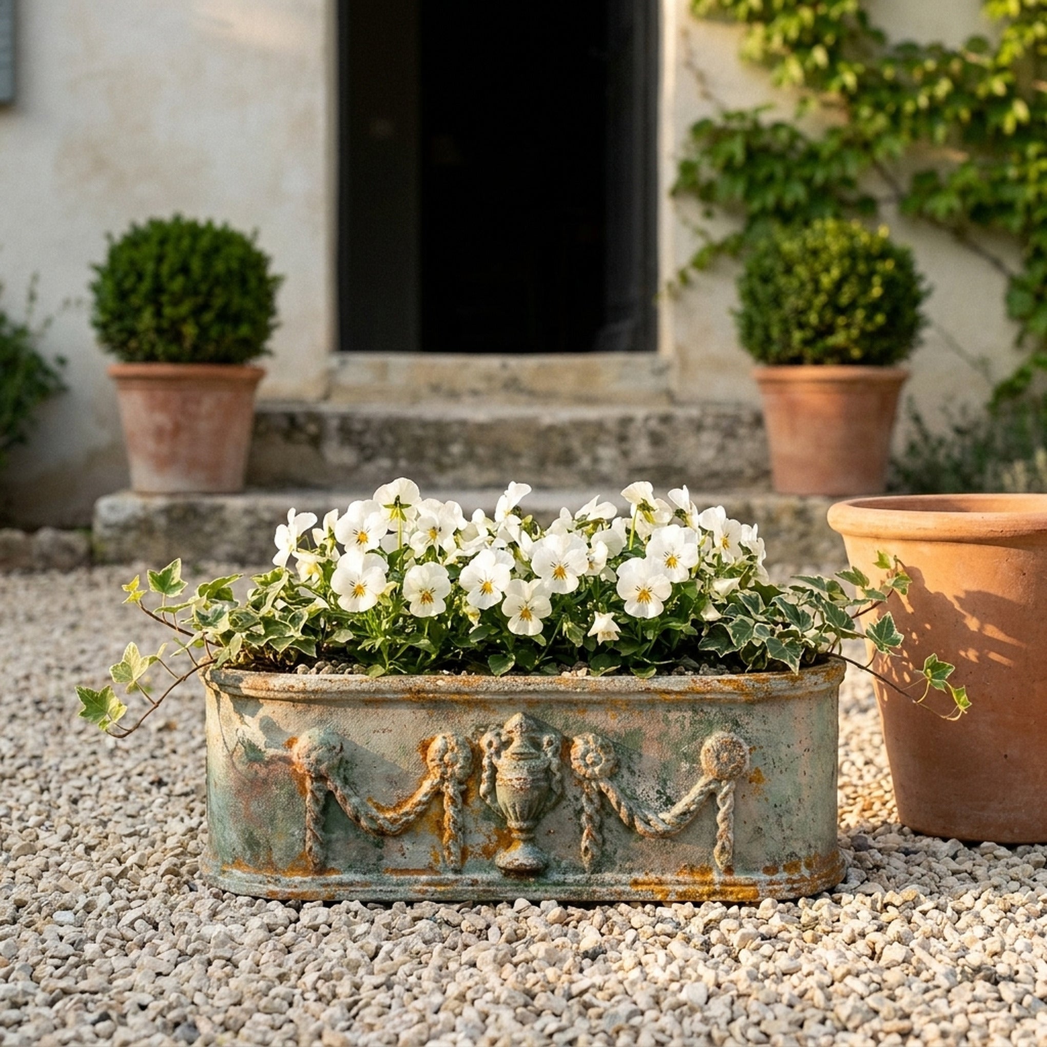 The Heritage Planter in weathered antique bronze cast iron planted with white violas and trailing ivy, on gravel in a provencal courtyard