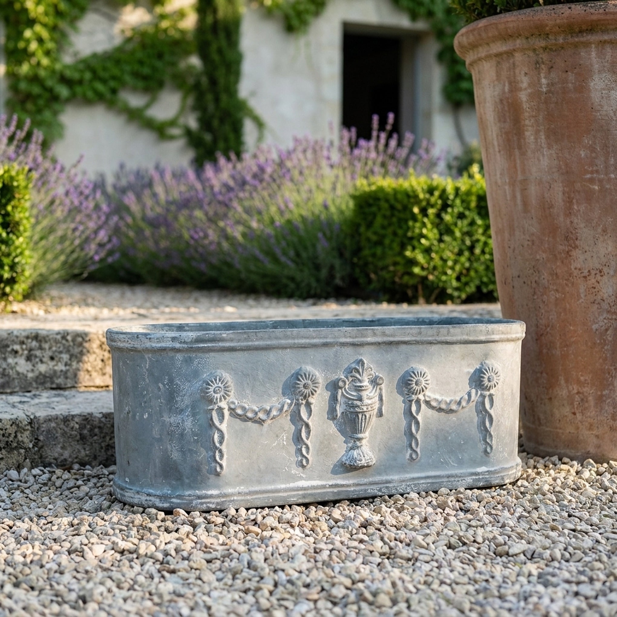 The Heritage Planter in weathered grey cast iron, an oval trough with swag relief, on gravel in a provencal courtyard