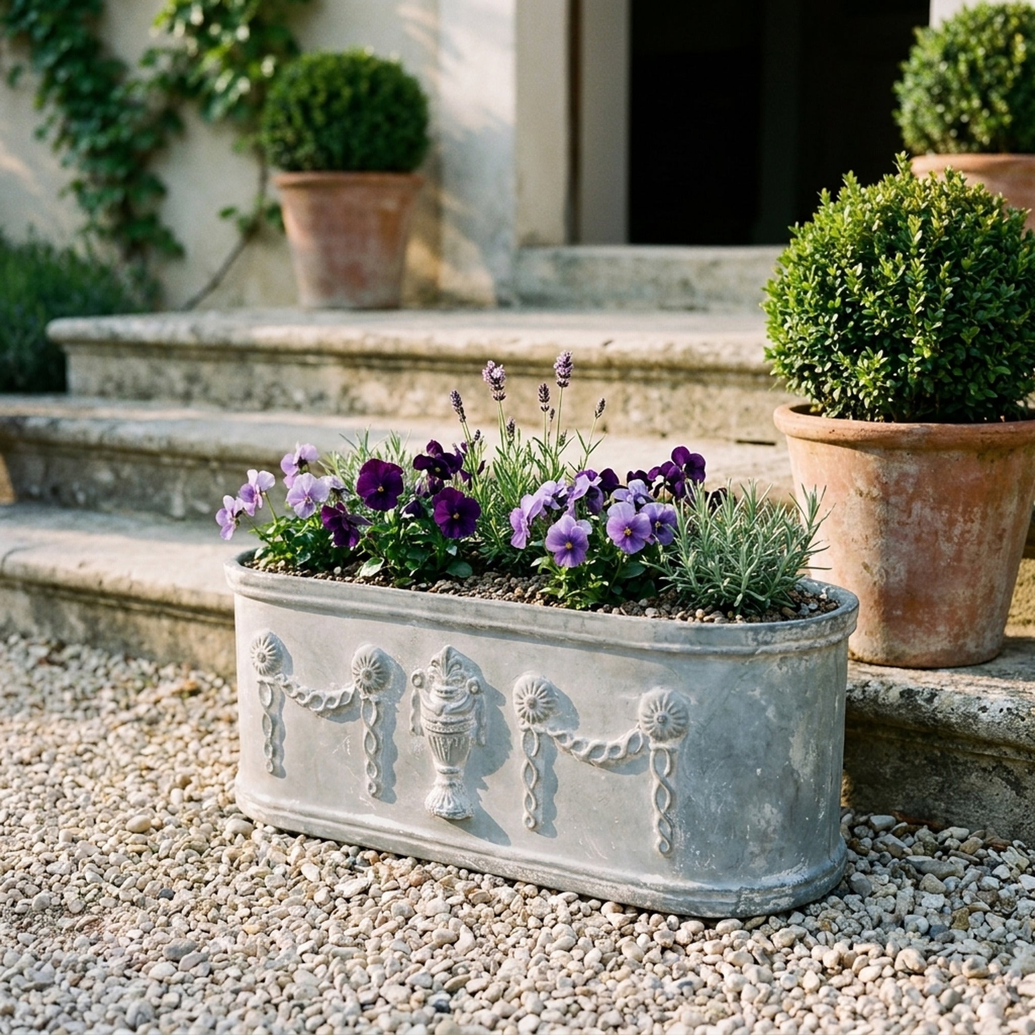 The Heritage Planter in weathered grey cast iron planted with purple and lavender violas, on gravel in a provencal courtyard