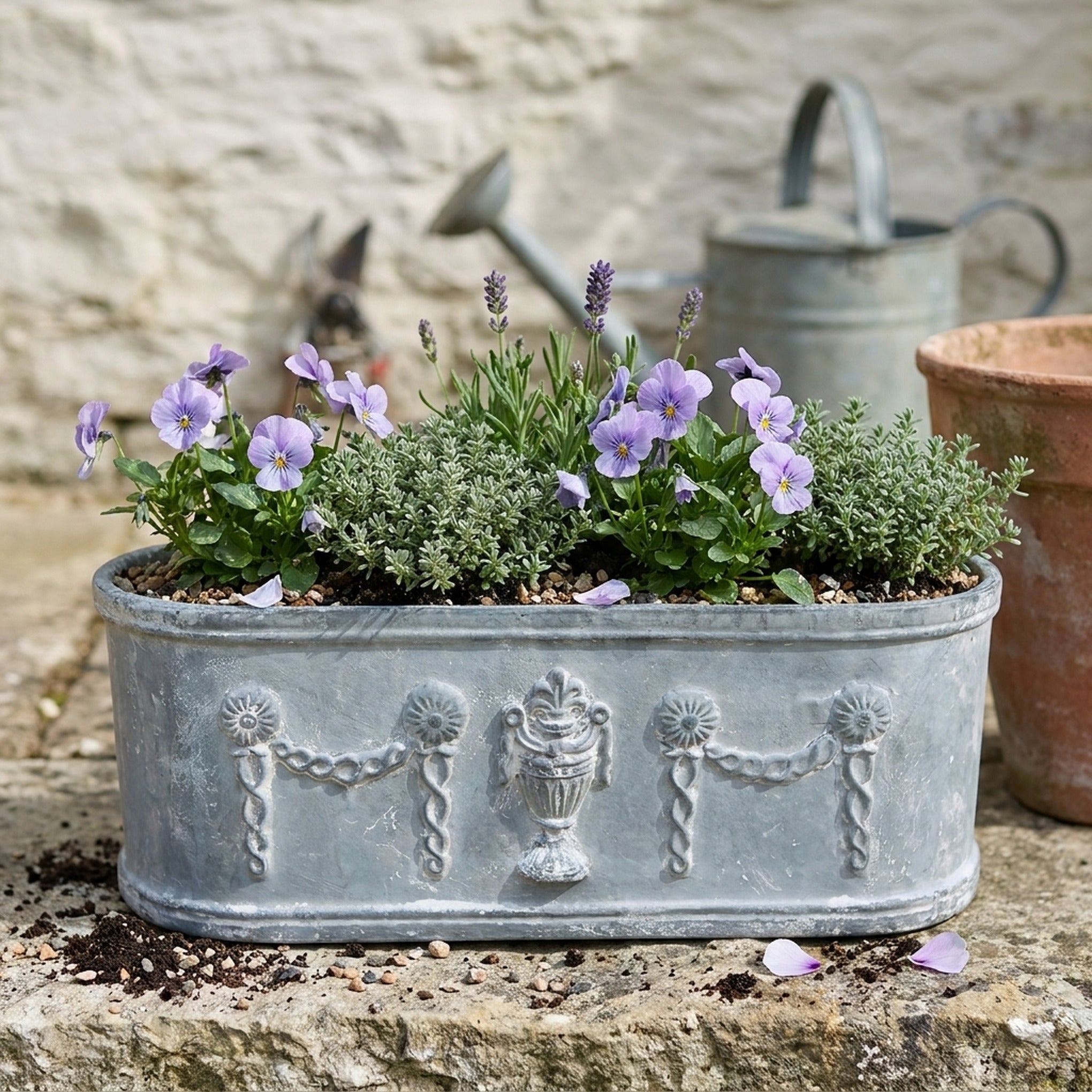 The Heritage Planter in weathered grey cast iron planted with lavender violas and thyme, side on along a limestone ledge