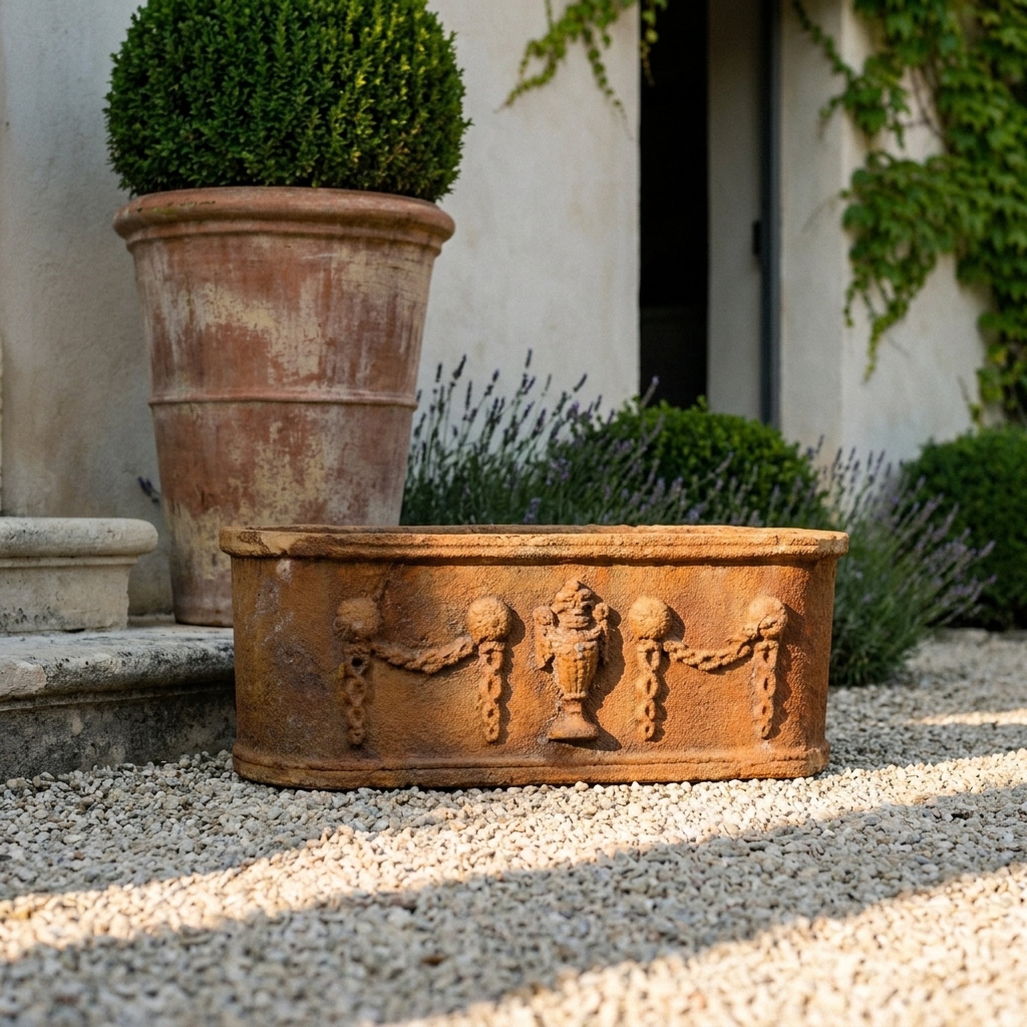 The Heritage Planter in rustic brown cast iron, an oval trough with swag relief, on gravel in a courtyard