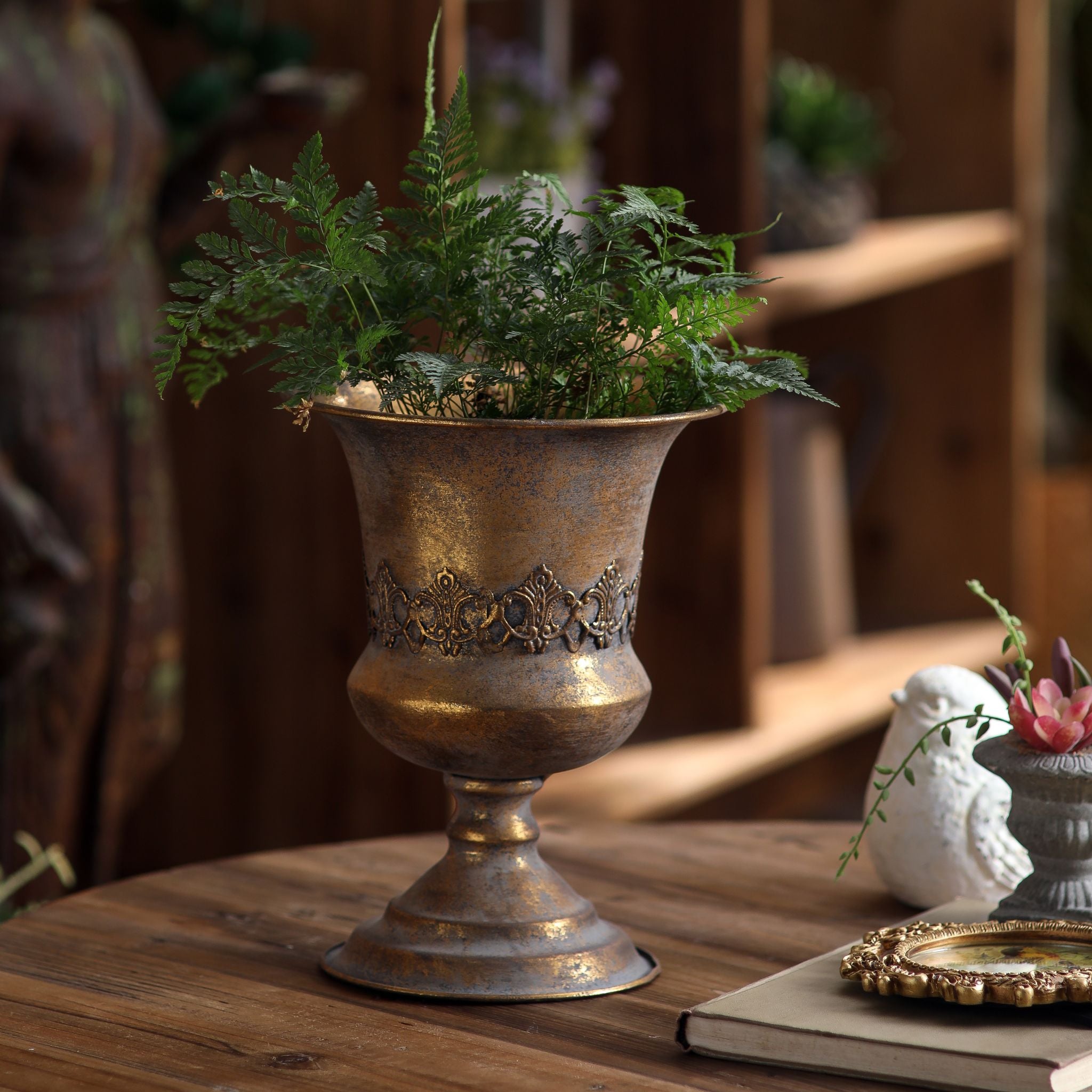 Magda French Provincial pedestal urn in antique gold holding a fern on a rustic timber table. Hero.