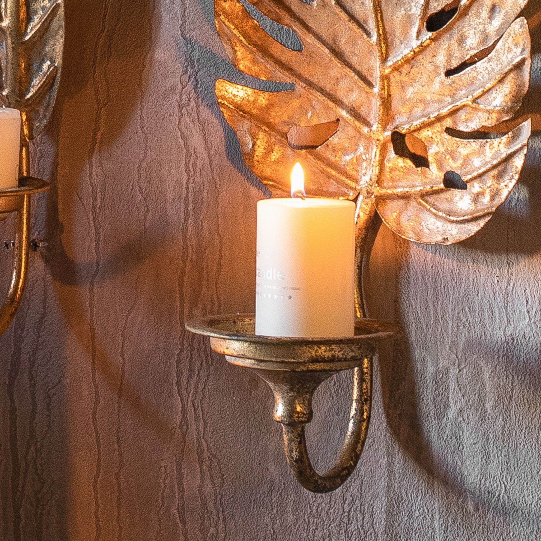 Lower half of the Adele wall sconce showing the candle cup and scrolled arm with a lit pillar candle below the split leaf.