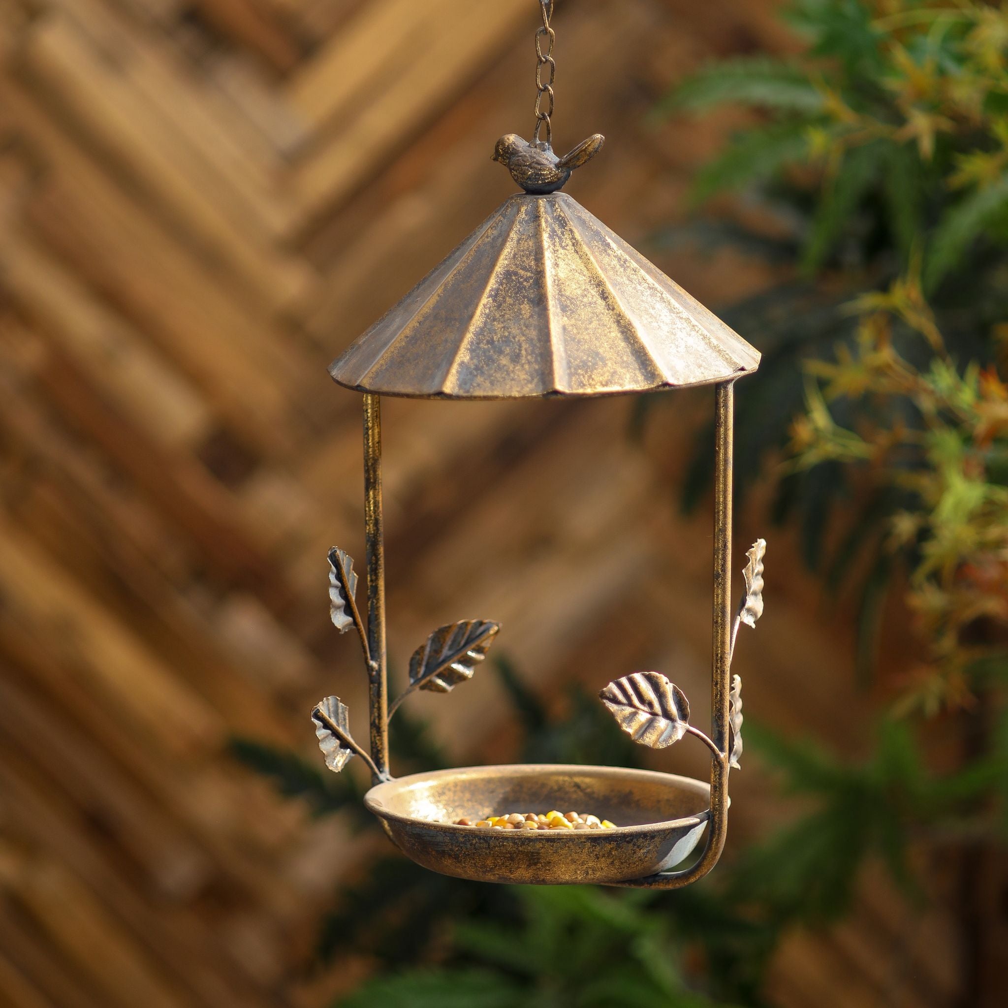 Nathalie antique gold hanging bird feeder hung outdoors against a timber fence and greenery.