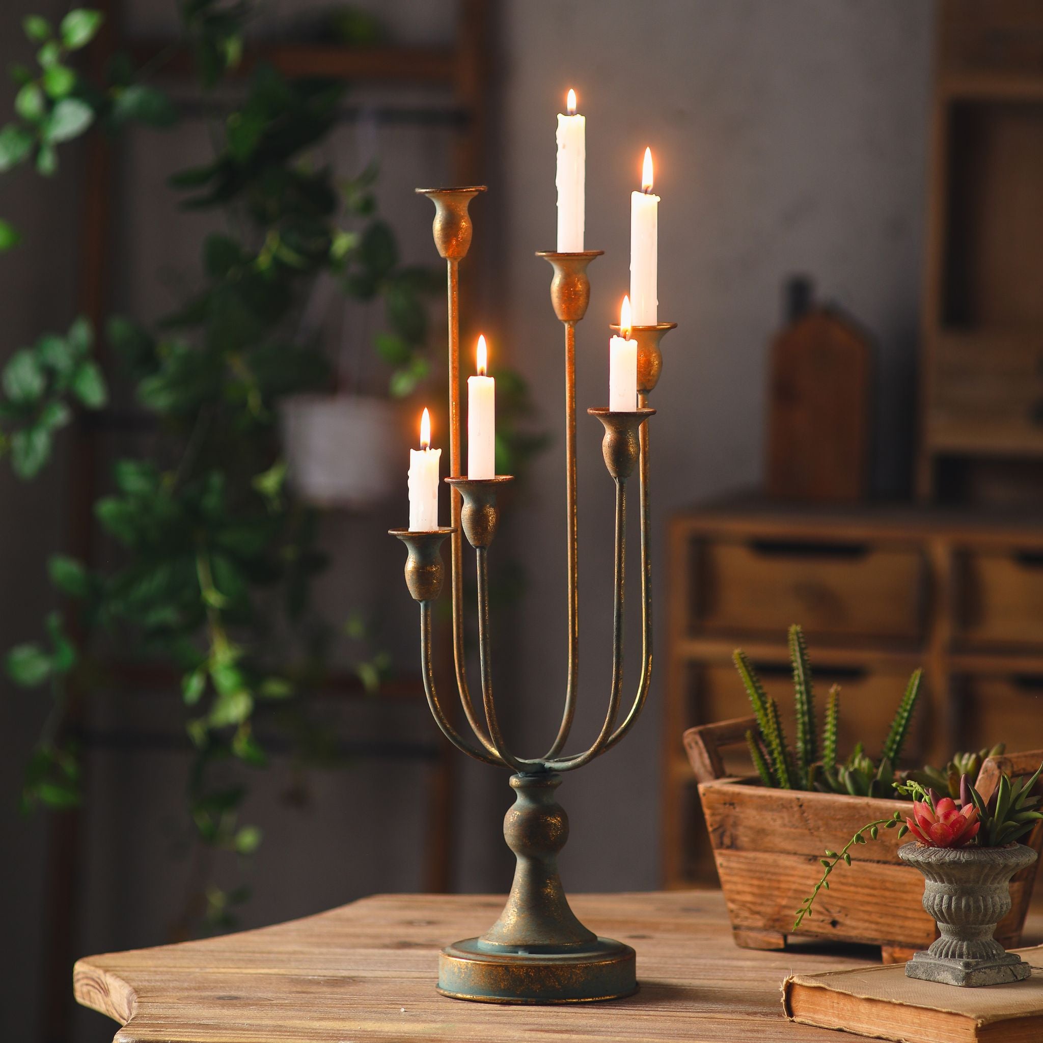 Audrée French Provincial table candelabra in antique gold aged metal, six trumpet cups holding lit taper candles, on a turned baluster base, styled on a timber table. Hero.