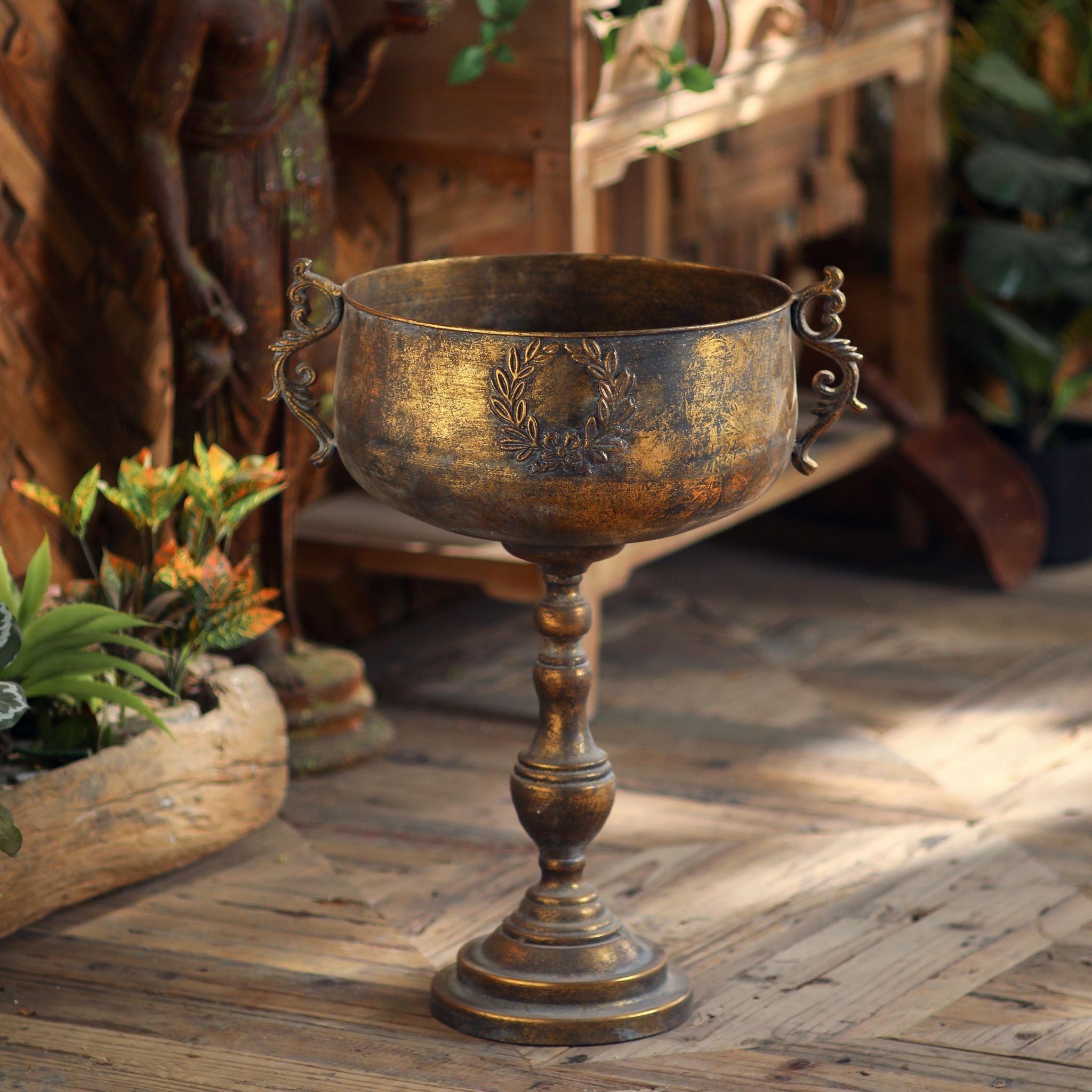 Empty antique gold footed urn planter showing the coupe bowl, laurel-wreath crest, scroll handles and turned pedestal foot