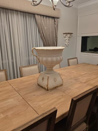 A large cream-colored decorative urn centerpiece placed on a light wood dining table in a dining room setting with upholstered chairs, curtains, and a chandelier.