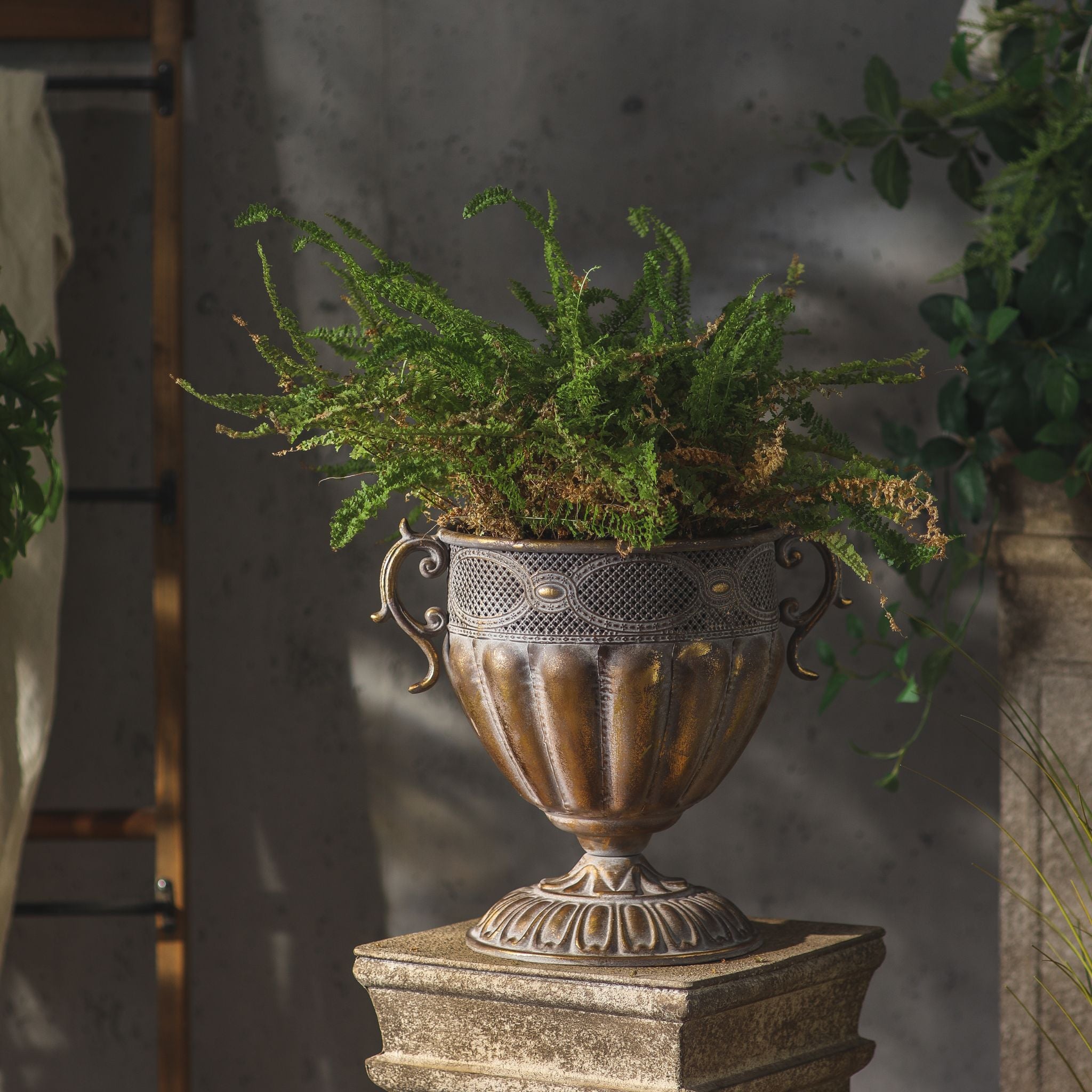 Benjamin French Provincial urn planter in antique gold metal, planted with a fern on a stone plinth. Hero.