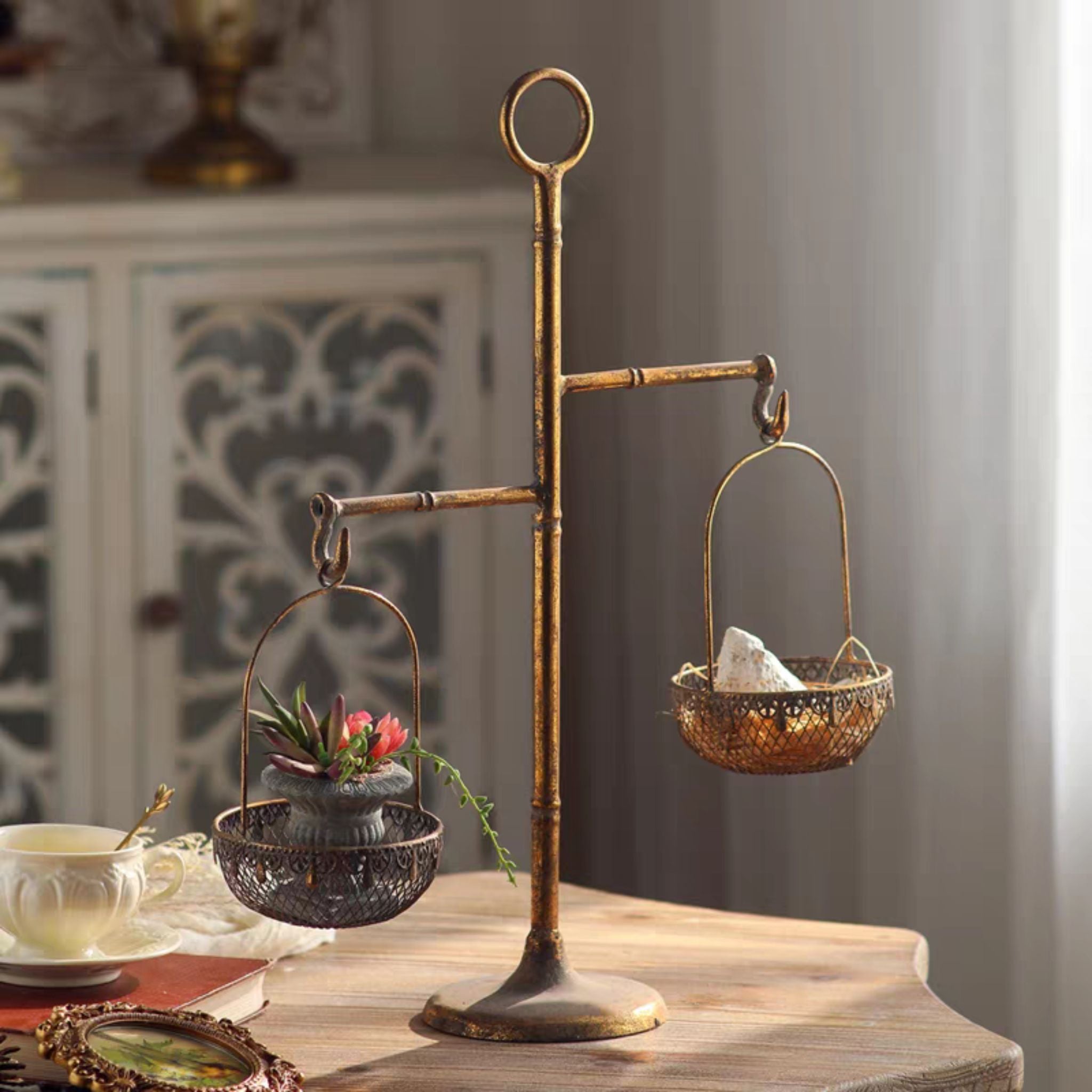 Bertrand French Provincial two tier basket stand in antique gold metal, styled on a wooden table with a potted succulent and crystals. Hero.