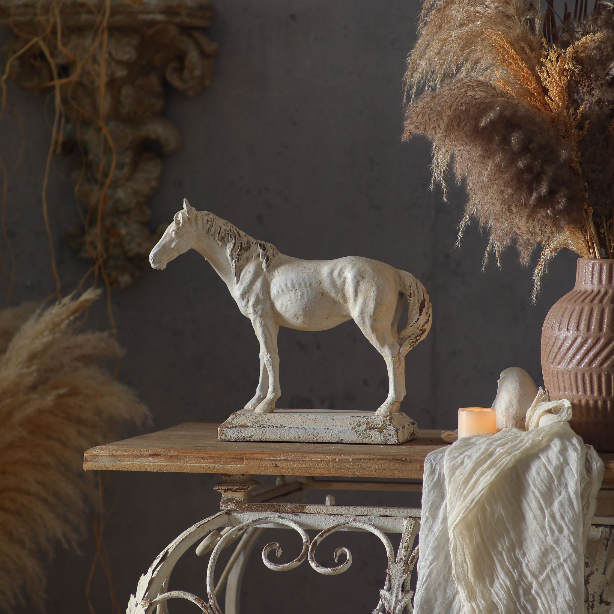 Camargue French Provincial horse sculpture in aged cream cast stone, styled on a vintage iron side table with pampas grass and a stone wall sconce behind. Hero.