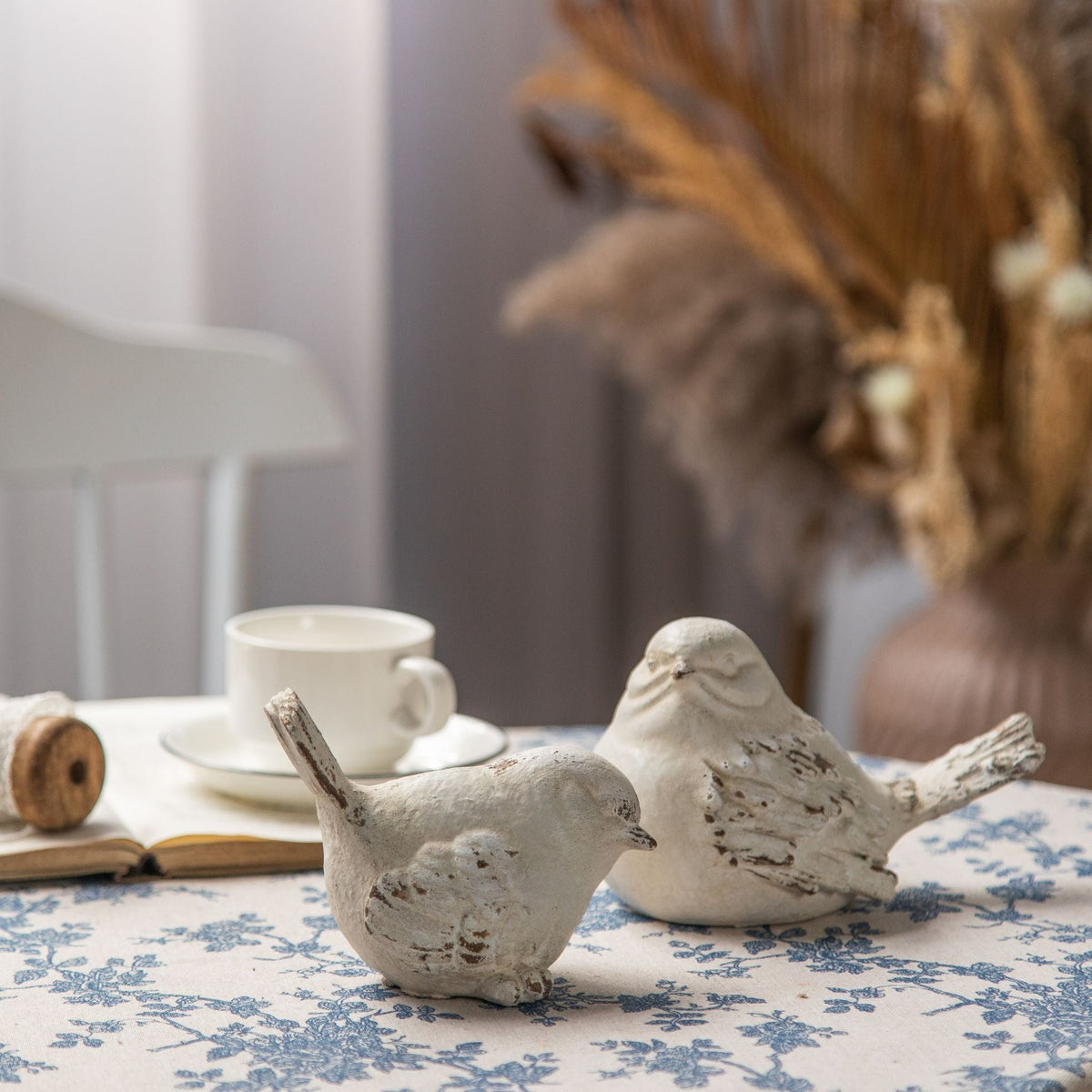 Cameron whitewashed bird figurines, set of two, on a blue toile cloth with a candlestick and an open book