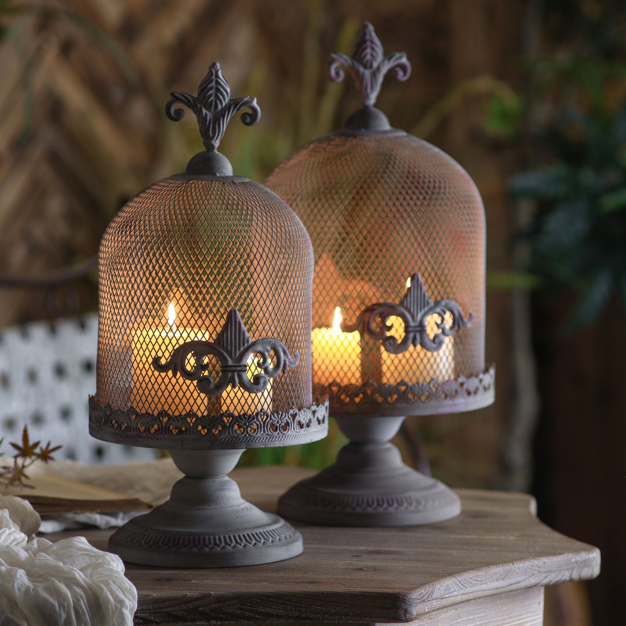 Cécile and Fleurine French Provincial candle cloches lit at dusk on a timber side table with timber lattice and garden grasses behind. Hero.