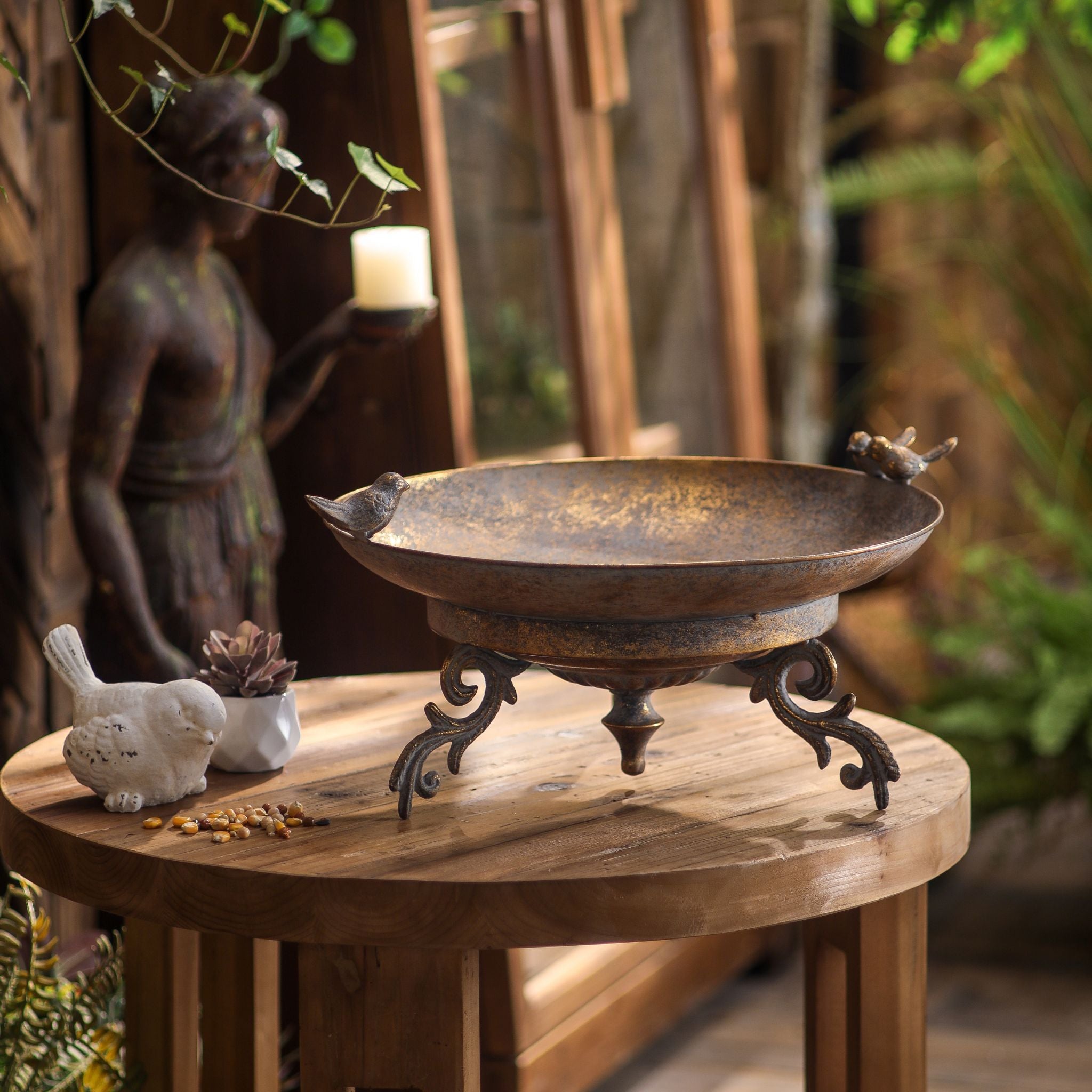 handler footed bird feeder dish in antique bronze metal, shallow bowl on scrolled feet with little perched bird figures, styled on a timber table. Hero