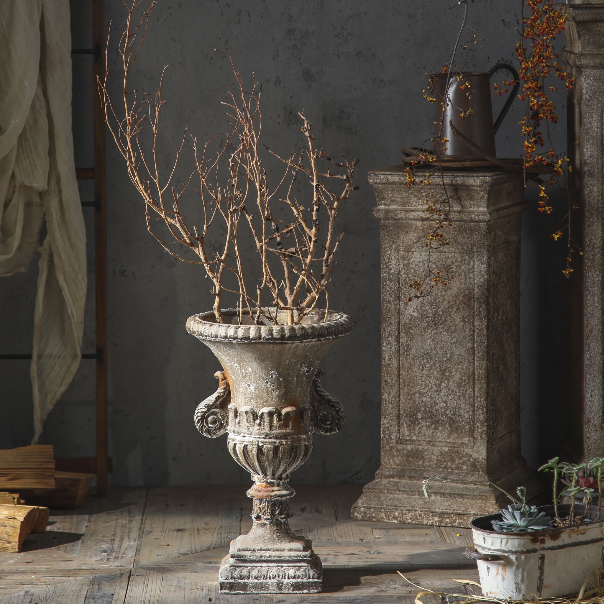 Emilie French Provincial scroll-handle garden urn planter in weathered patina, planted with bare branches. Hero.