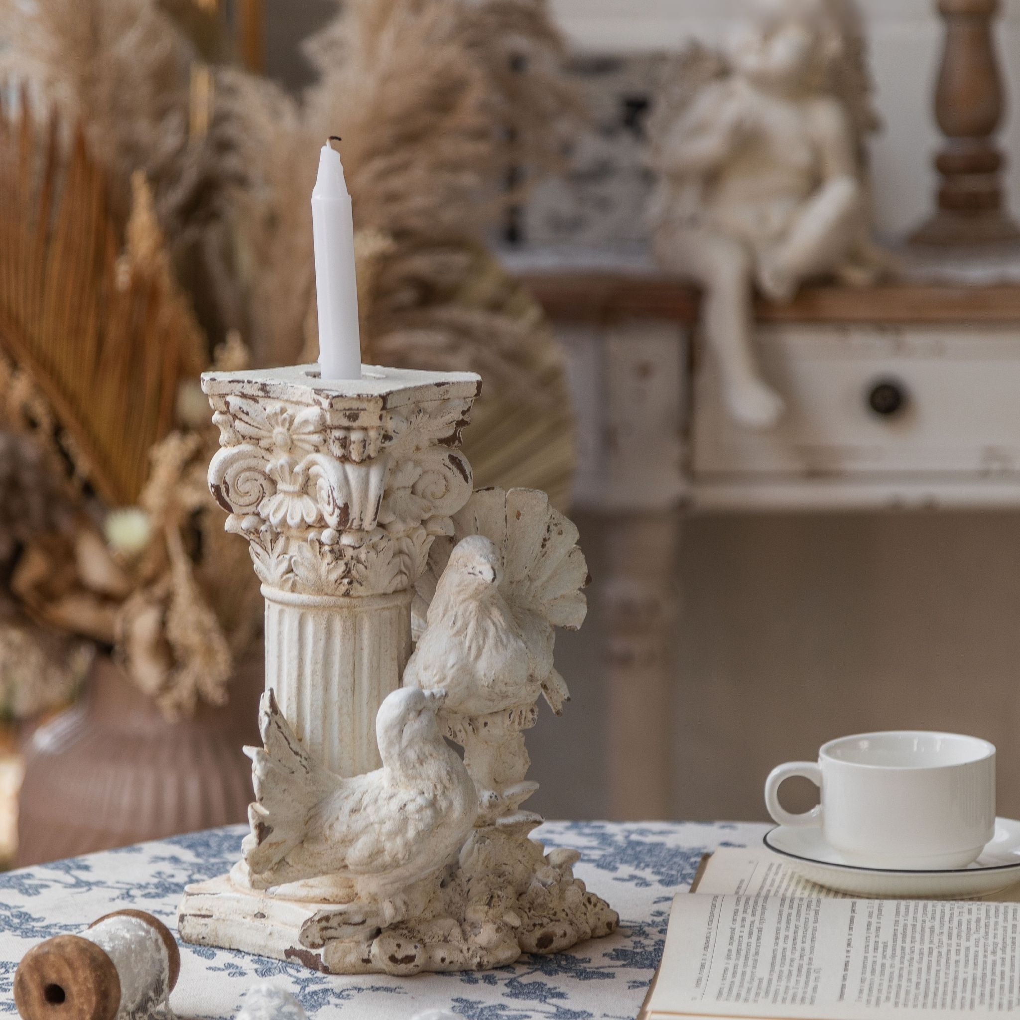 Étienne French Provincial dove candle holder in distressed antique white, fluted column with two doves at the base and a lit taper, styled on a table. Hero.