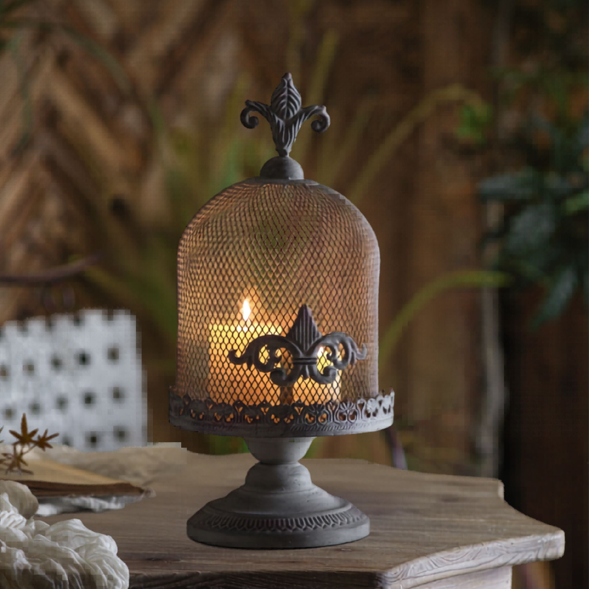 Fleurine French Provincial candle cloche close-up showing aged iron mesh dome, fleur-de-lis finial and scrollwork over a lit pillar candle. Hero.