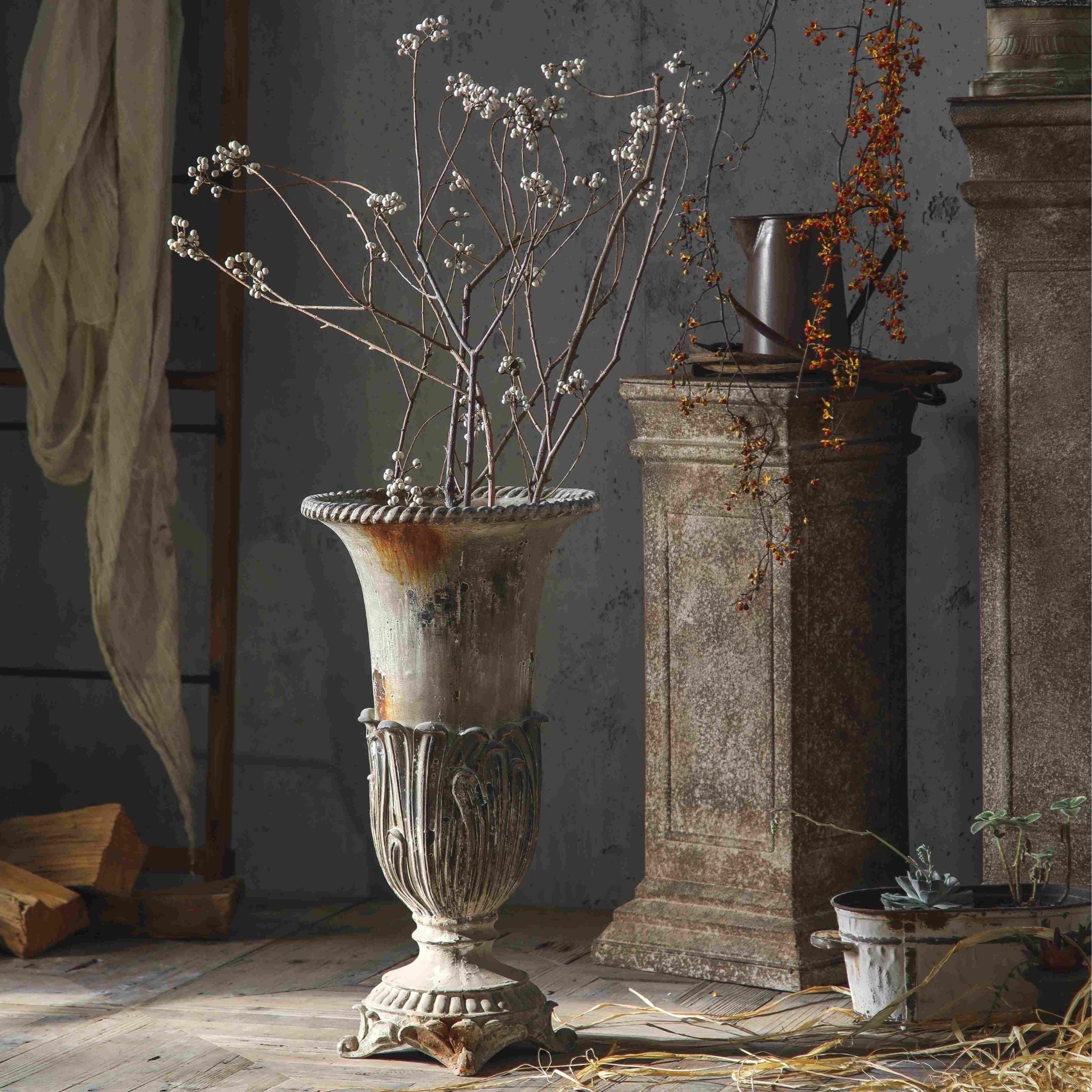 Florent urn vase styled with branches beside a stone plinth and aged pitcher.