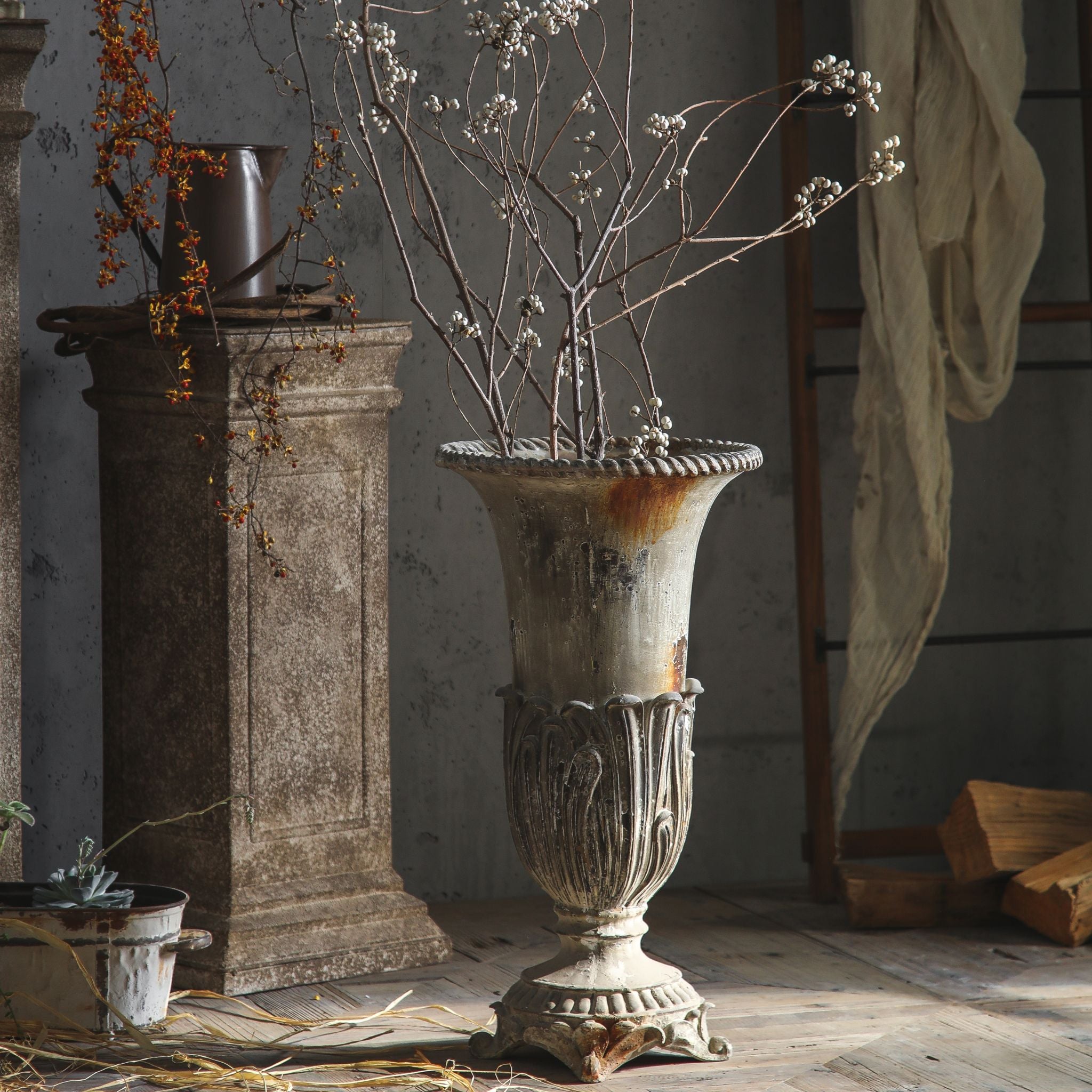 Florent tall campana urn vase in weathered grey-white, holding white-berry branches, in a stone-and-timber room. Hero.