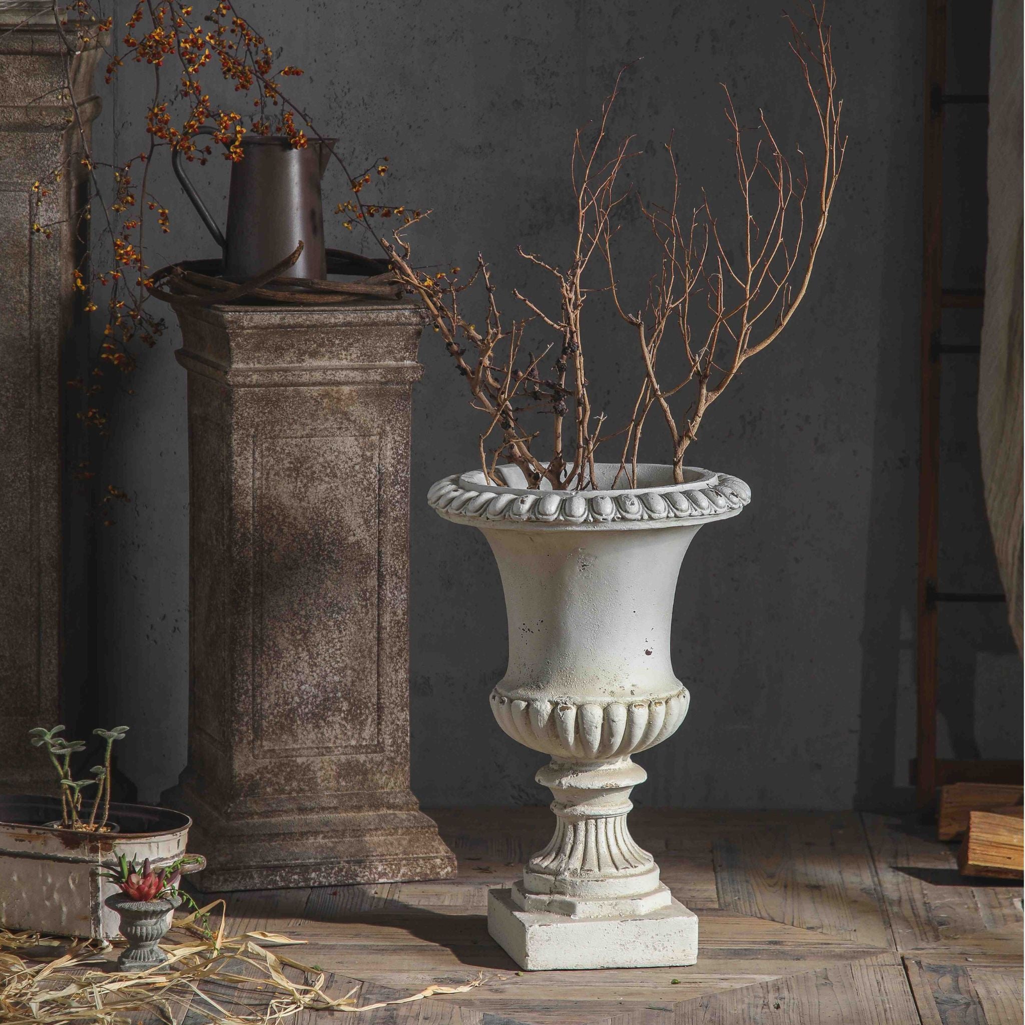 Florentine French Provincial Medici urn planter in distressed antique white, styled with bare branches on timber flooring. Hero.