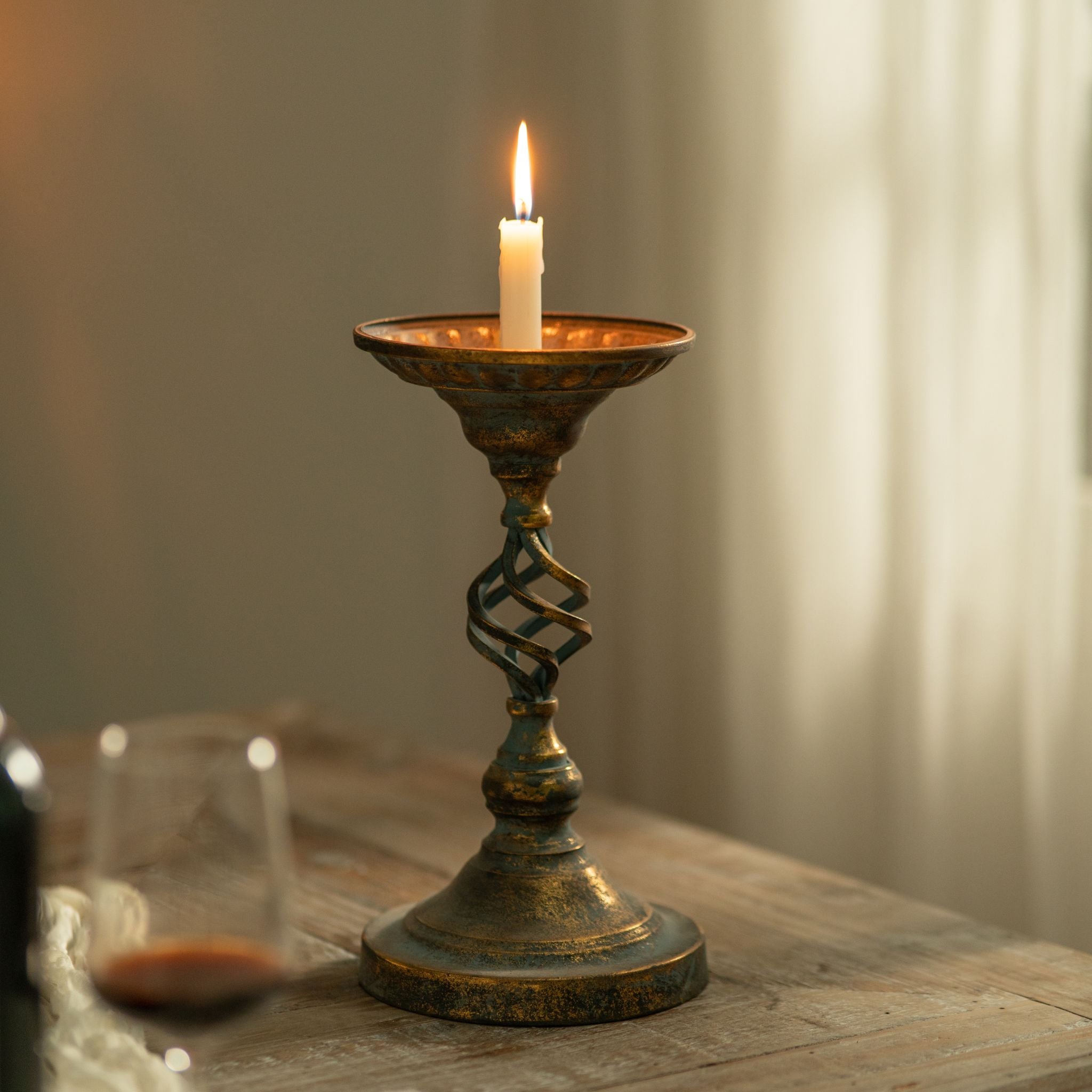 Francis aged-metal candle holder full front view, dished top and turned baluster foot, lit taper by a window.