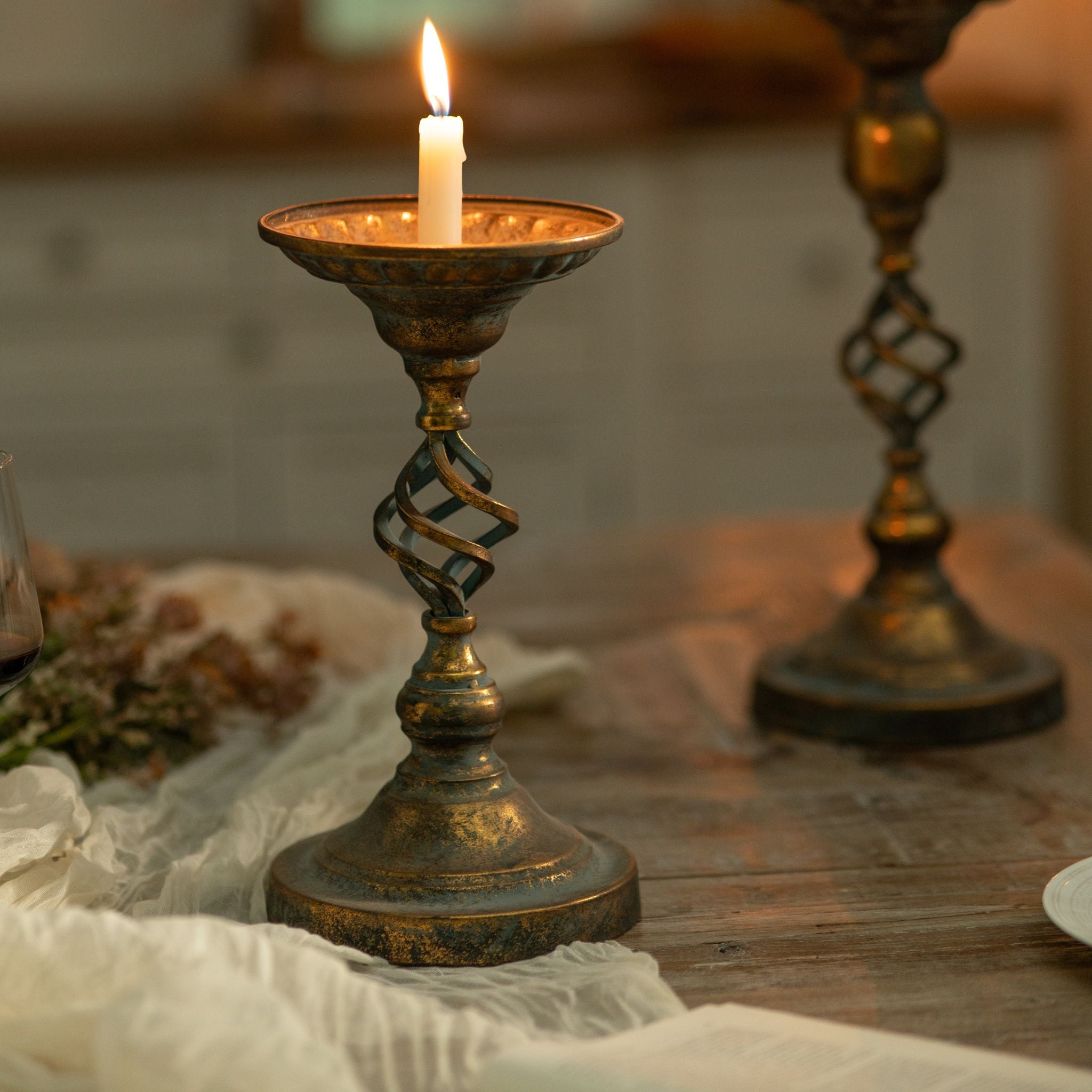 Francis French Provincial candle holder in antique gold and verdigris, twisted cage stem, lit taper, styled on a wooden dining table with the taller Michel behind. Hero.