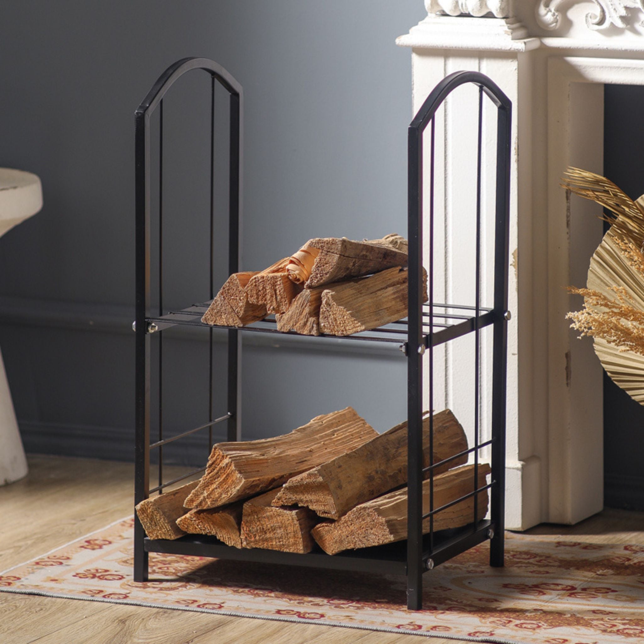 Didier two-tier matte black firewood log rack loaded with split logs and kindling beside a fireplace.