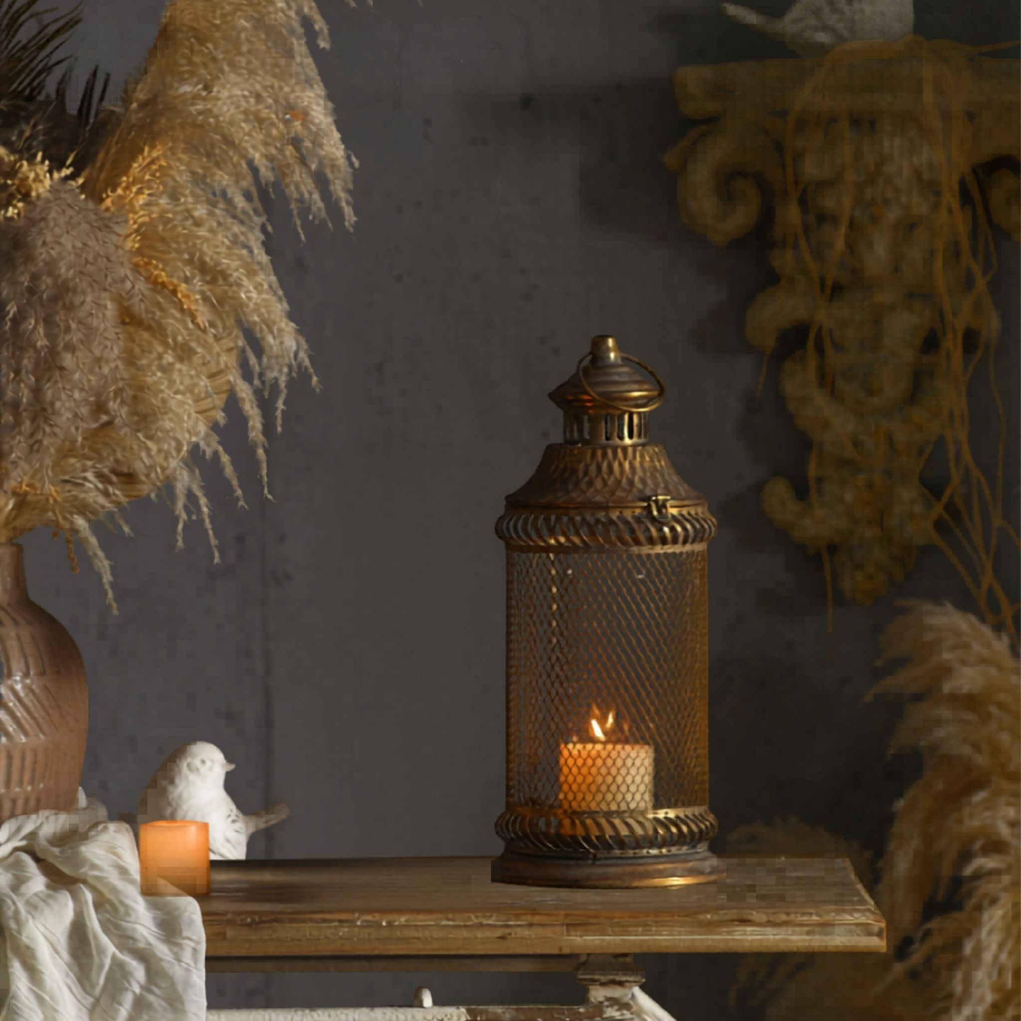 Arielle French Provincial candle lantern in antique gold aged metal, diamond mesh over a glass sleeve, lit candle inside, styled on a timber console with pampas grass.