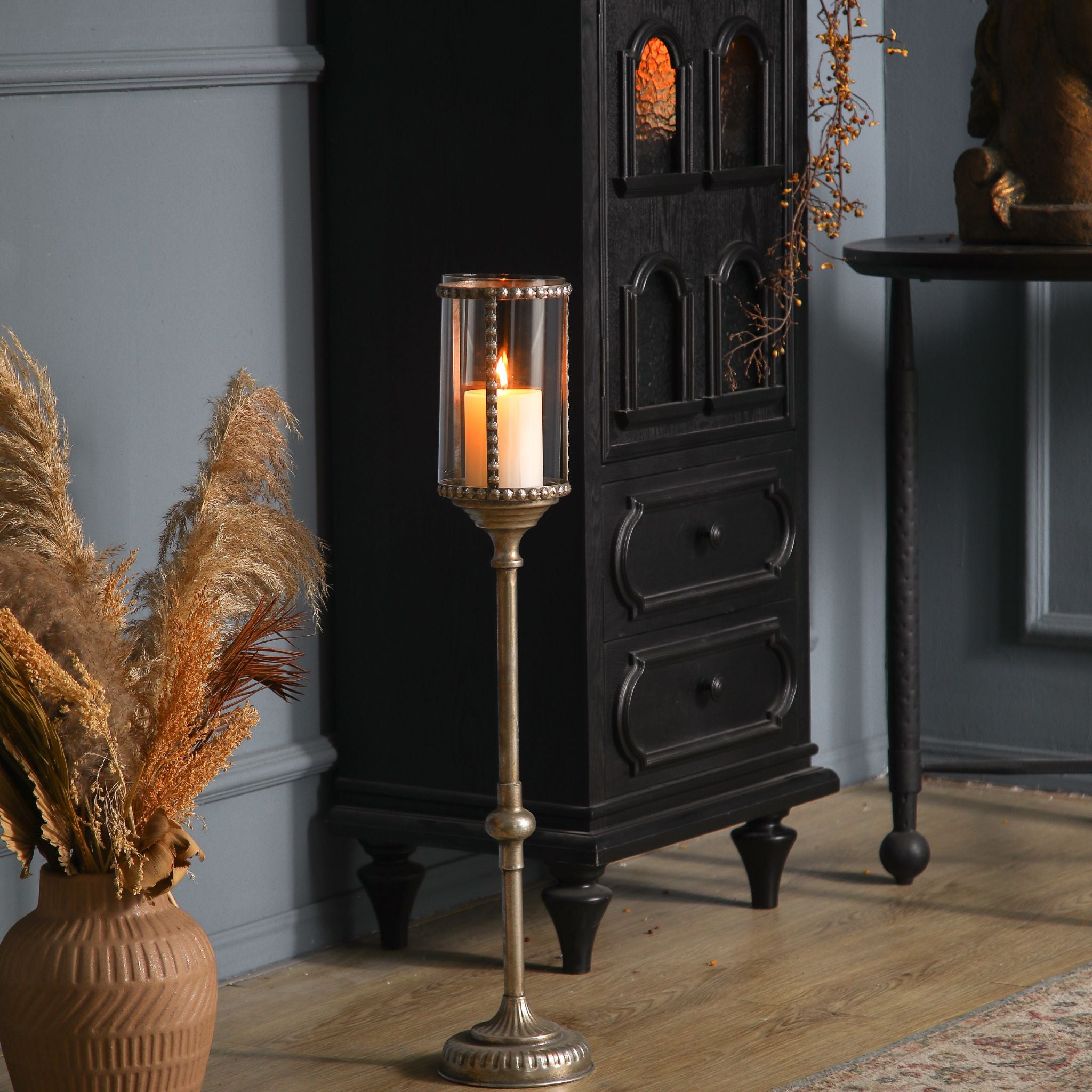 Sylvestre French Provincial floor candle holder in antique gold with a beaded glass hurricane and lit pillar candle, on a timber floor beside a black cabinet.