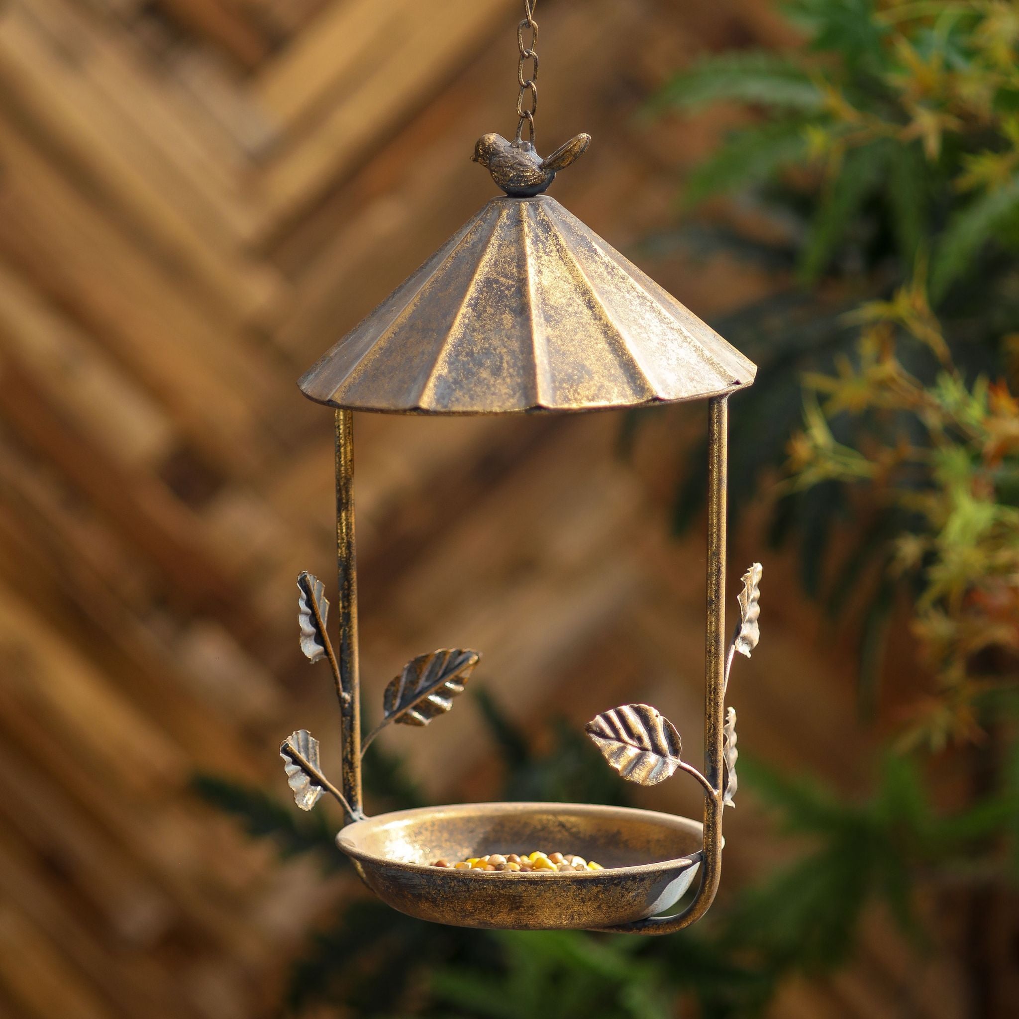 Nathalie antique gold hanging bird feeder with a fluted roof, leaf stems, and a little bird finial, seed in the dish.