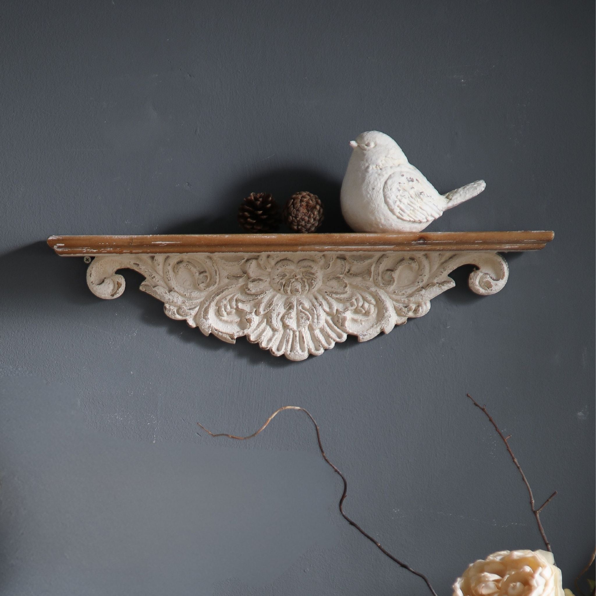 Jennifer French Provincial wall ledge shelf in distressed whitewash with carved floral apron and timber top, mounted upper right on a charcoal wall and styled with a small white bird and pinecones. Hero.