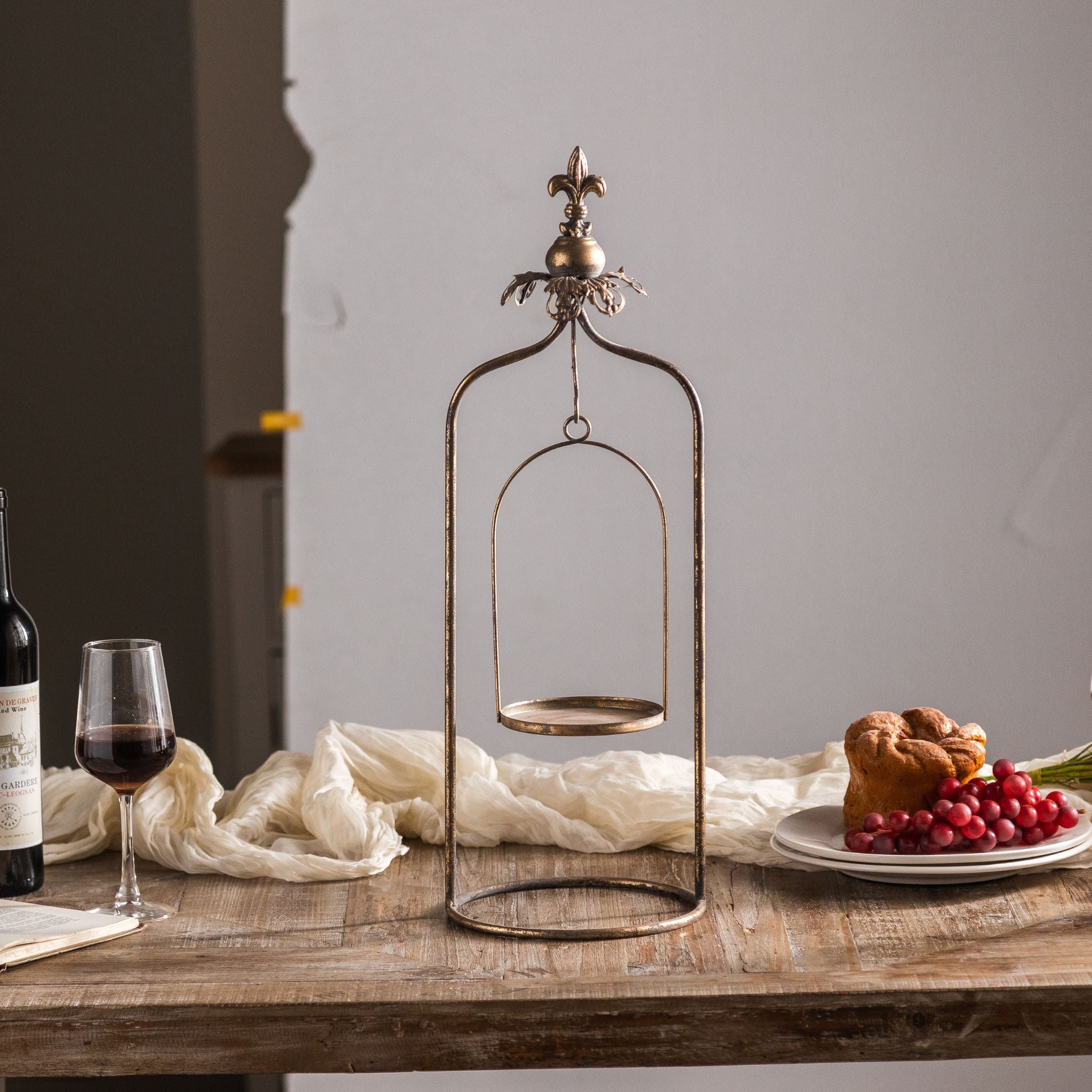 Julien aged-metal candle holder shown empty, full arched frame with fleur-de-lis finial, swinging cradle and round base.