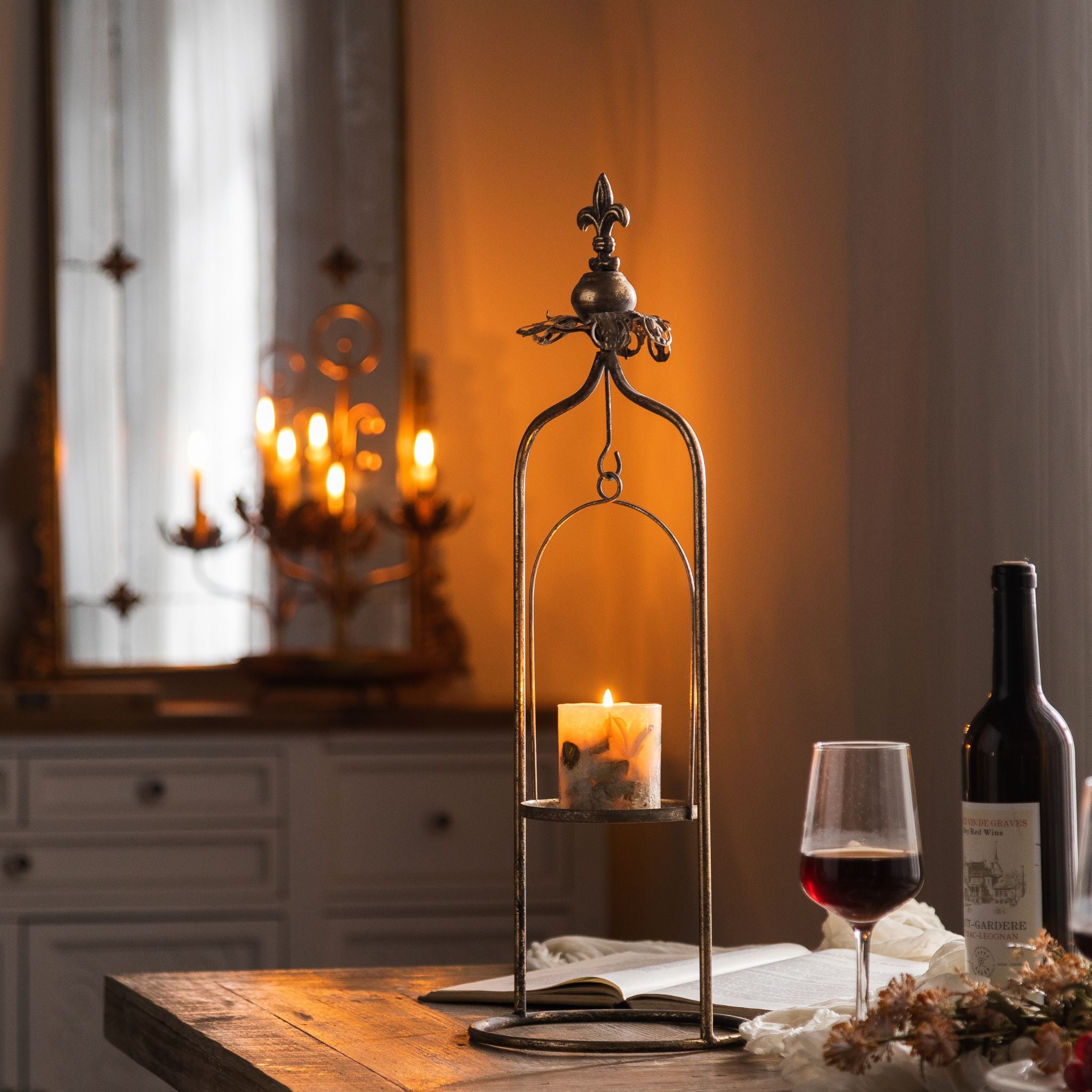 Julien French Provincial hanging candle holder in aged metal with a fleur-de-lis finial, lit pillar candle in the swinging cradle, styled on a dining table. Hero.