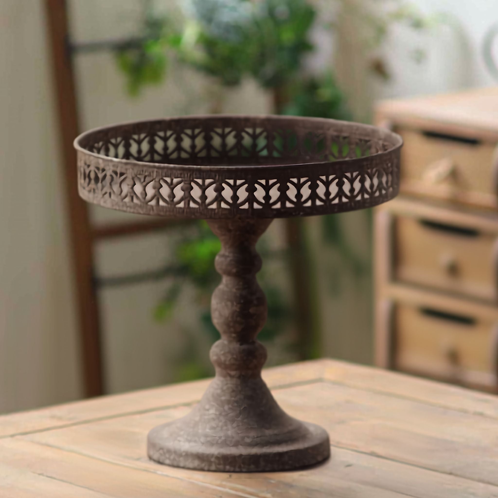 Lina French Provincial pedestal display stand in rustic rust-brown metal with a pierced gallery rim, on a timber table. Hero.