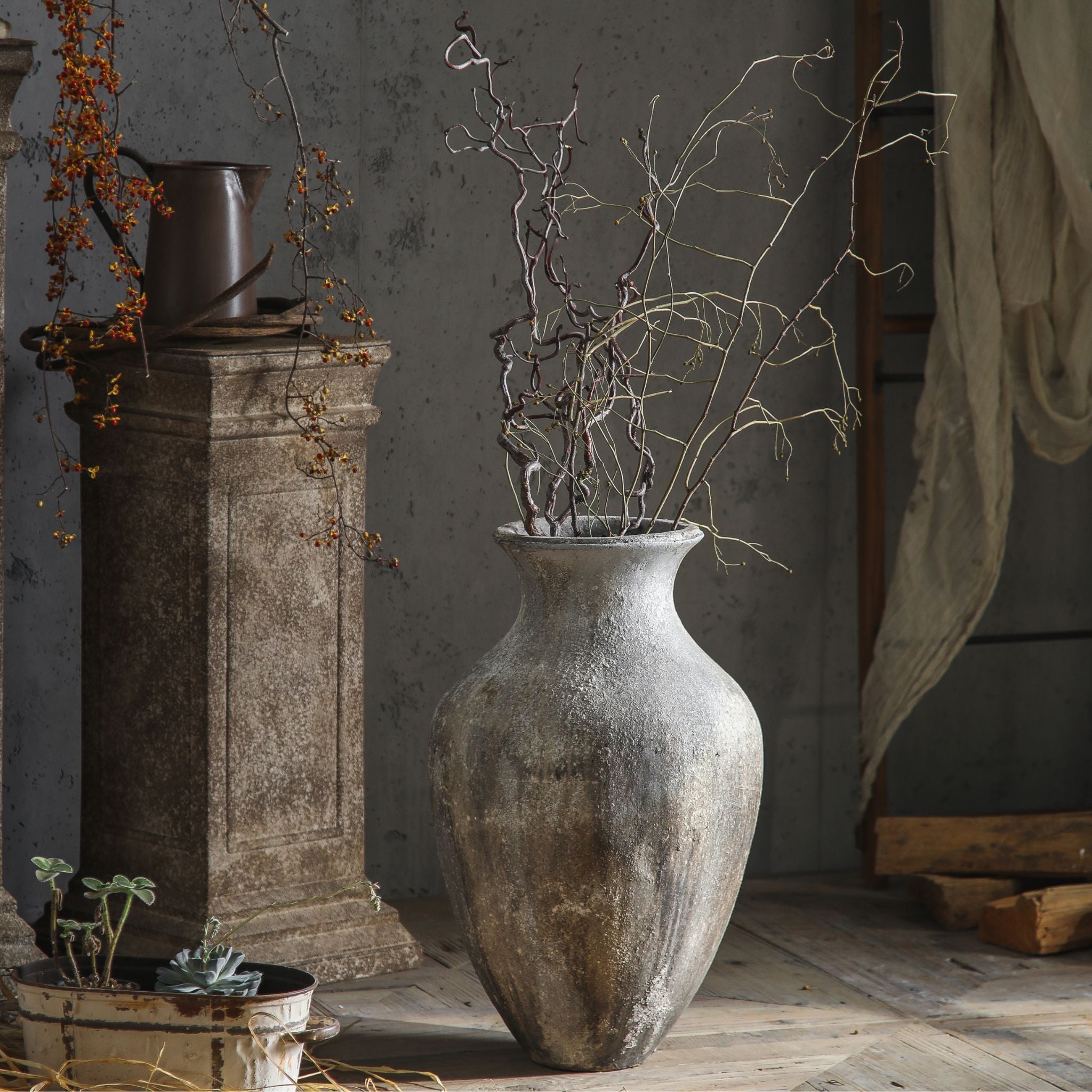 Lionel aged stone floor vase with curly willow branches beside a stone pedestal. Hero