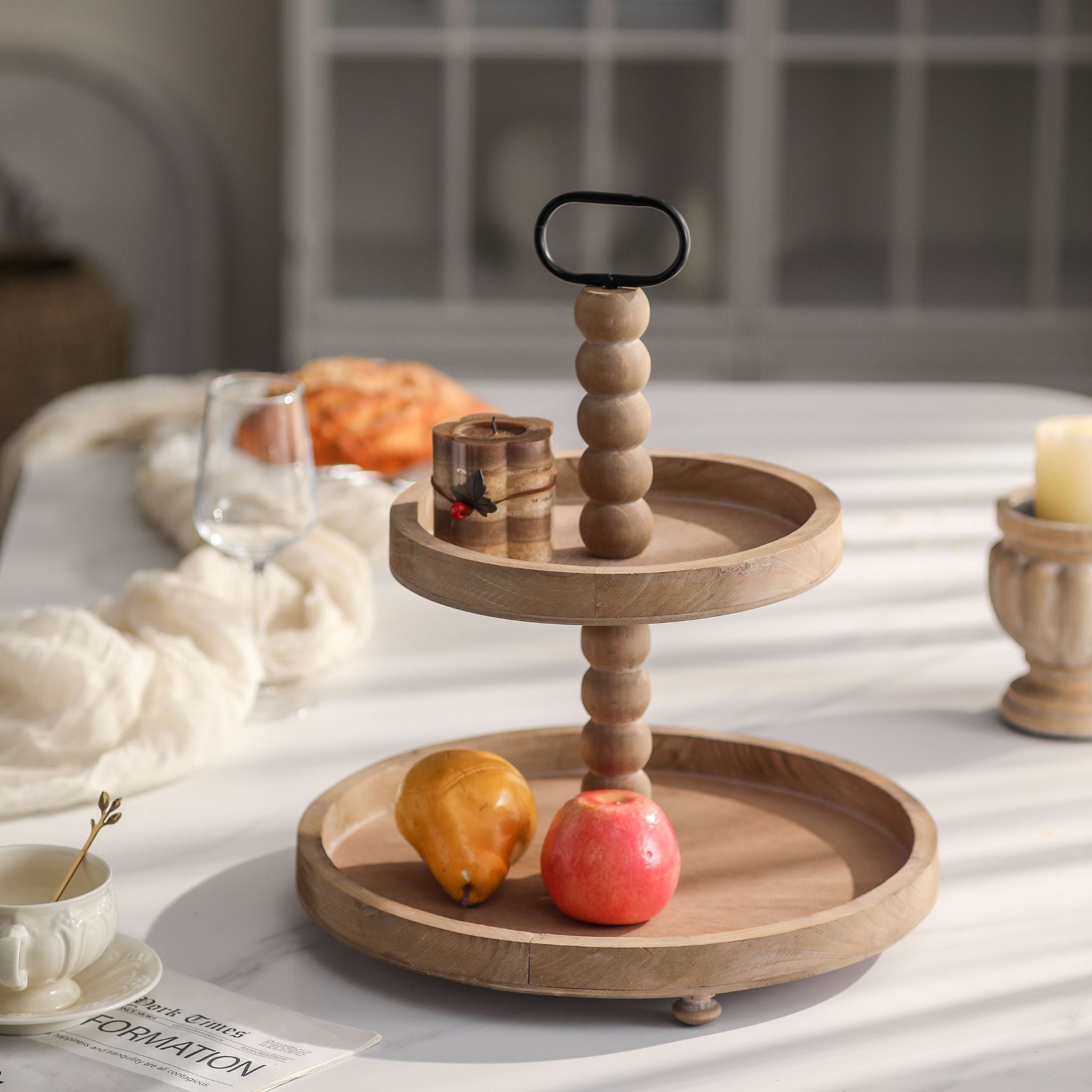 Mallory French Provincial two tier wooden tray stand in natural timber with a beaded stem and black metal handle, on a table. Hero.