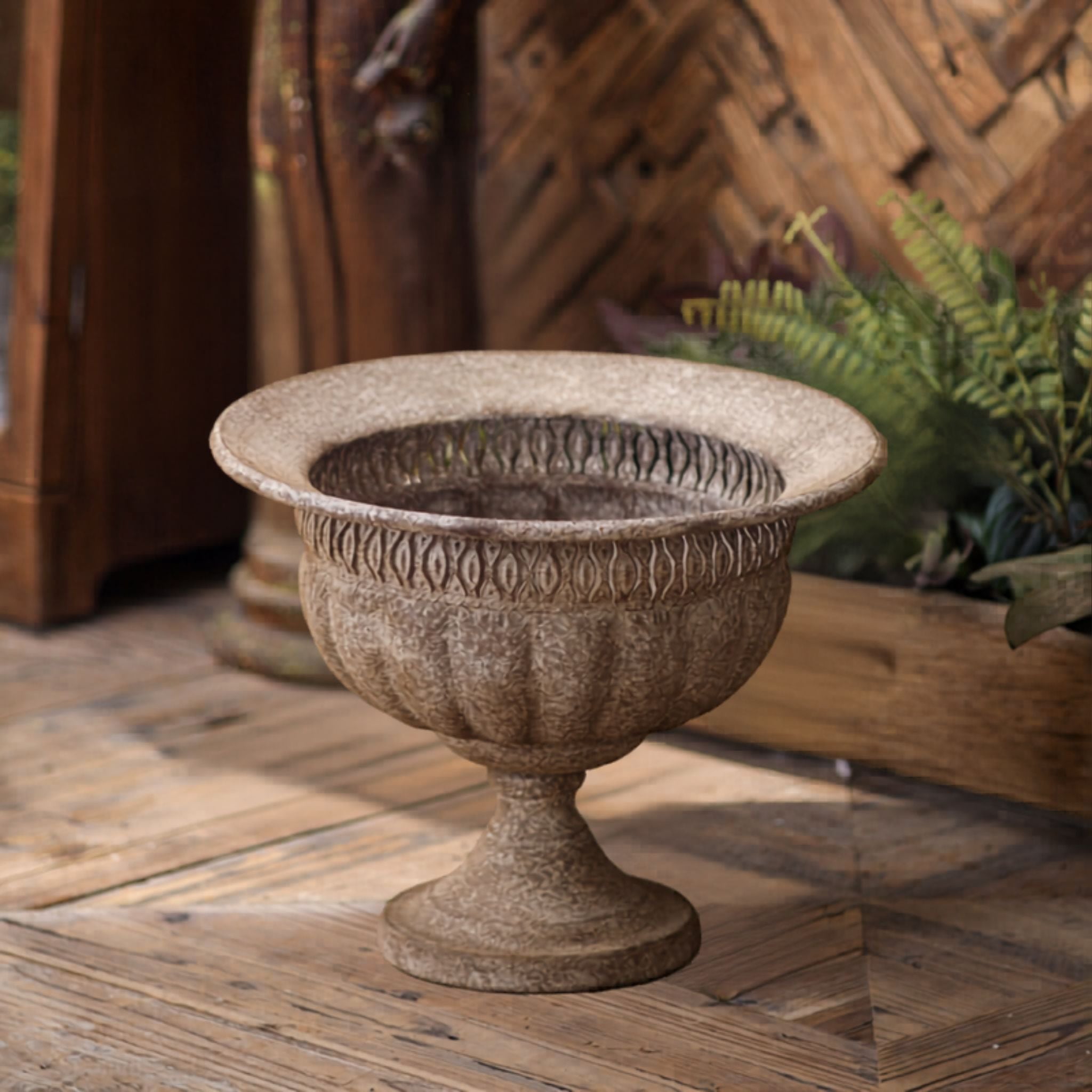 Myriam French Provincial footed urn planter in aged stone-look metal, styled on timber flooring beside a fern. Hero.