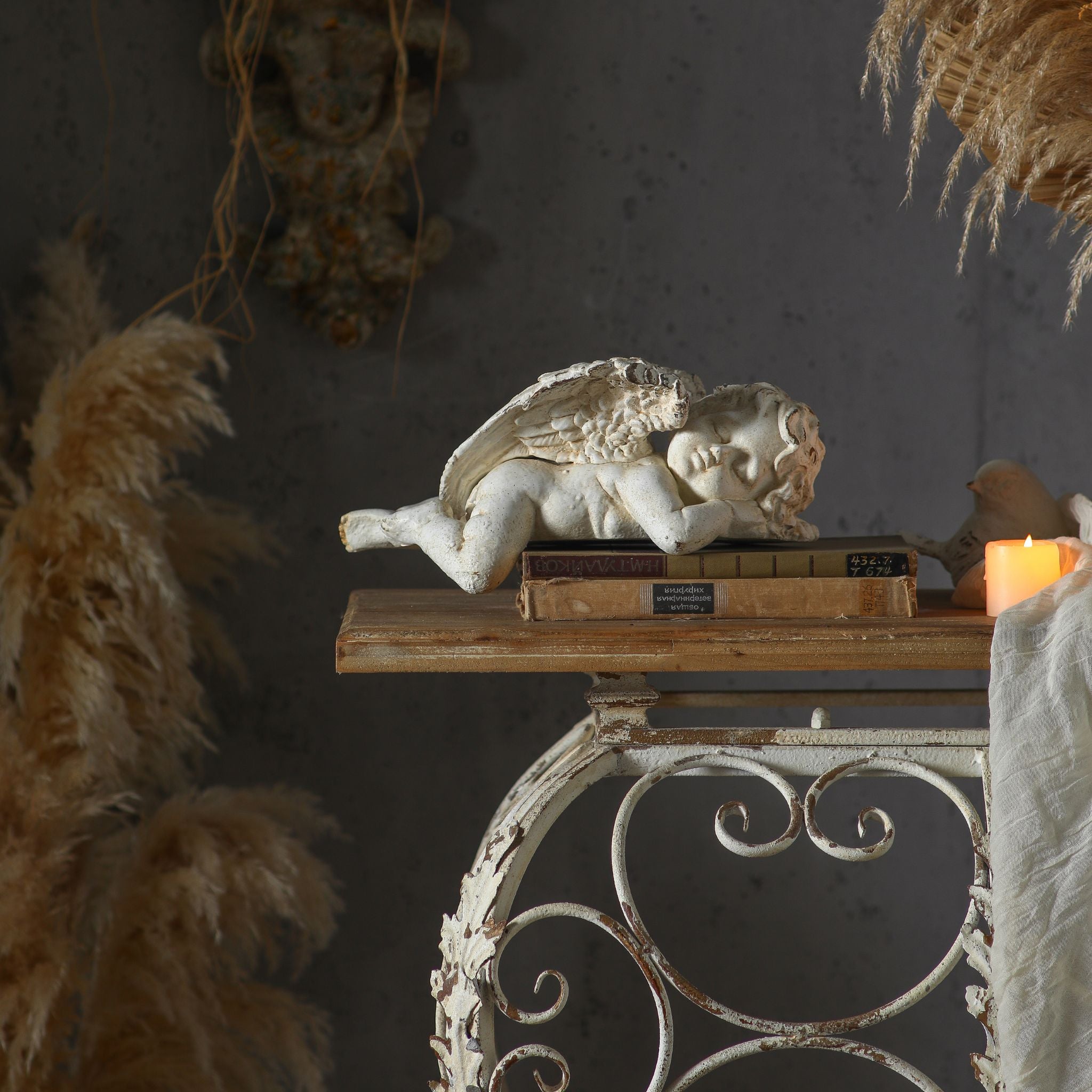 Wider styled vignette of Océane sleeping cherub on a wood and iron console with pampas grass, dried botanicals, and antique books for a French country dressing room