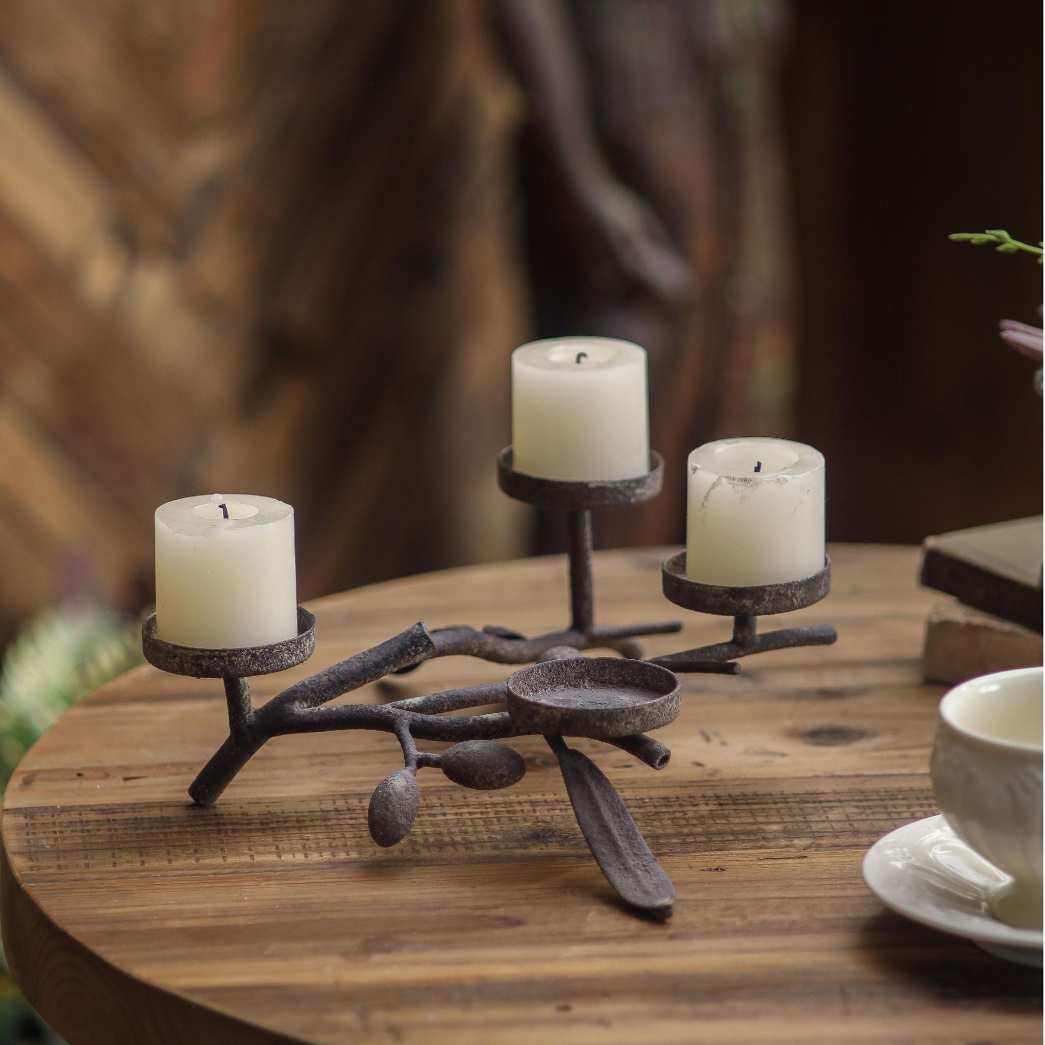 Marcus aged metal olive branch candle holder with three pillar candles on a wooden table. 