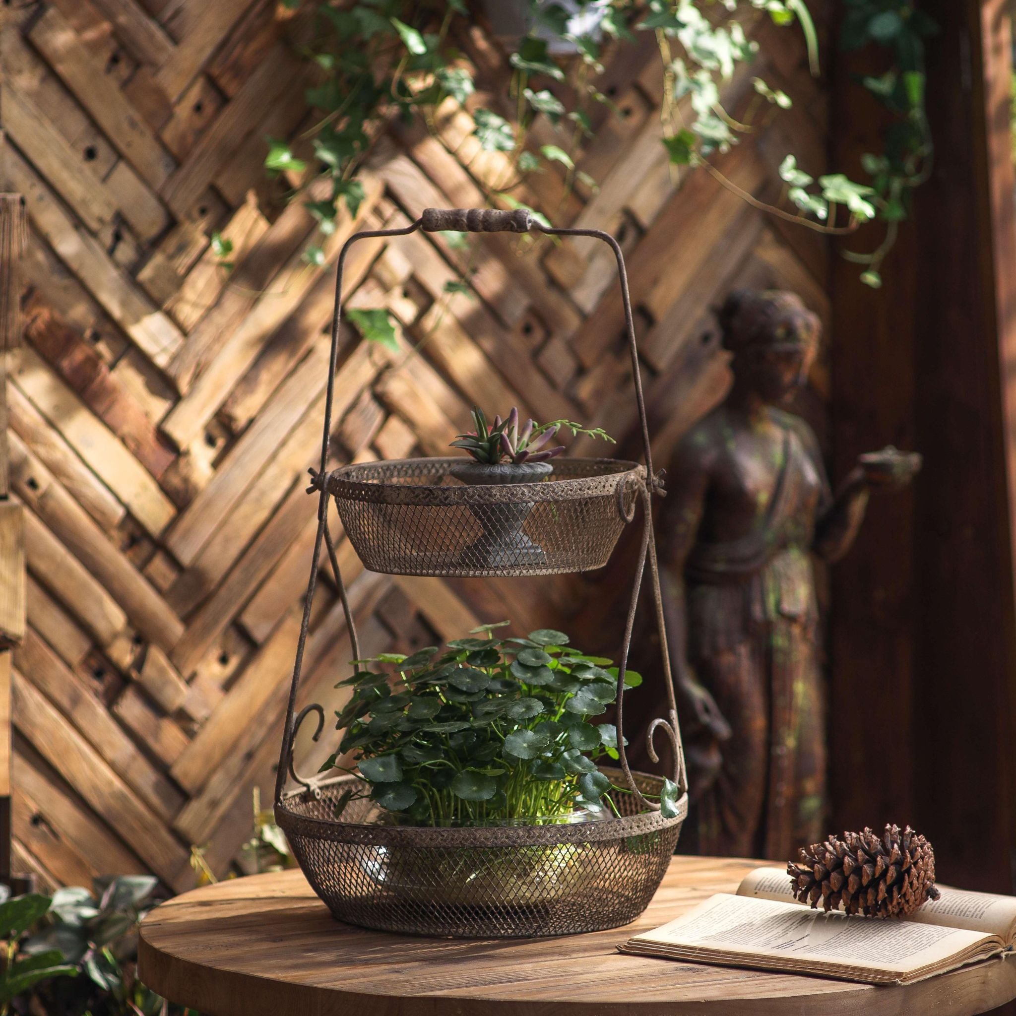 Oscar two tier basket carrier in rustic rust-brown mesh metal, two graduated baskets on a scrolled frame with a grip handle, styled with a planted lower basket. Hero.