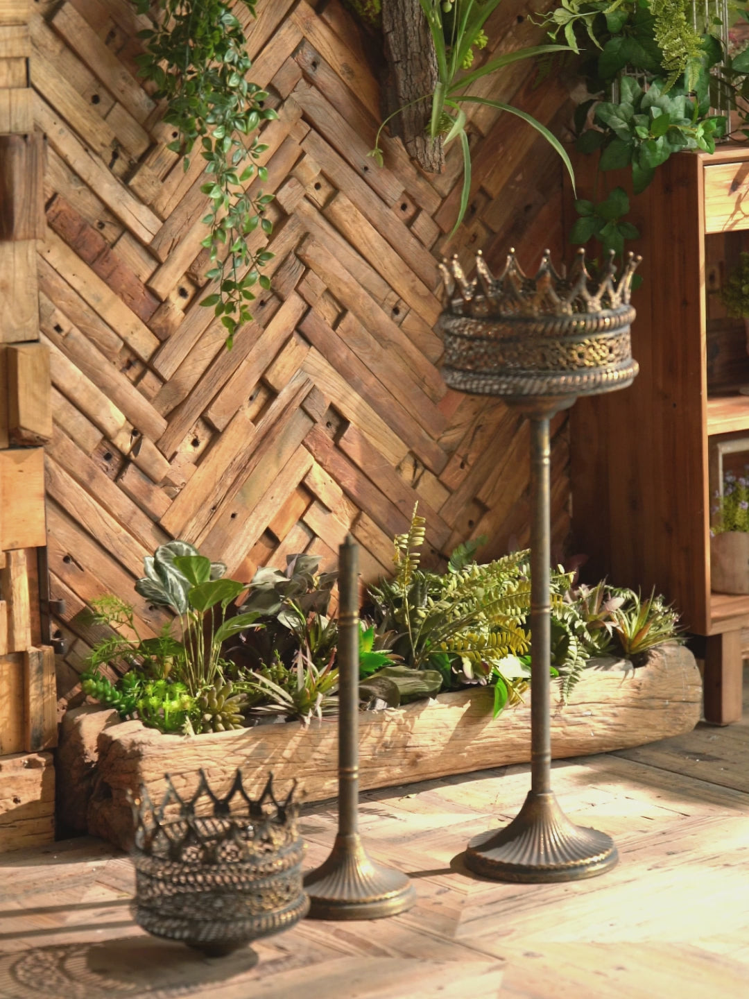 Solange crown-topped pedestal planter in aged gold and verdigris metal, styled in a rustic room with a column and trailing greenery, shown with its taller companion planted