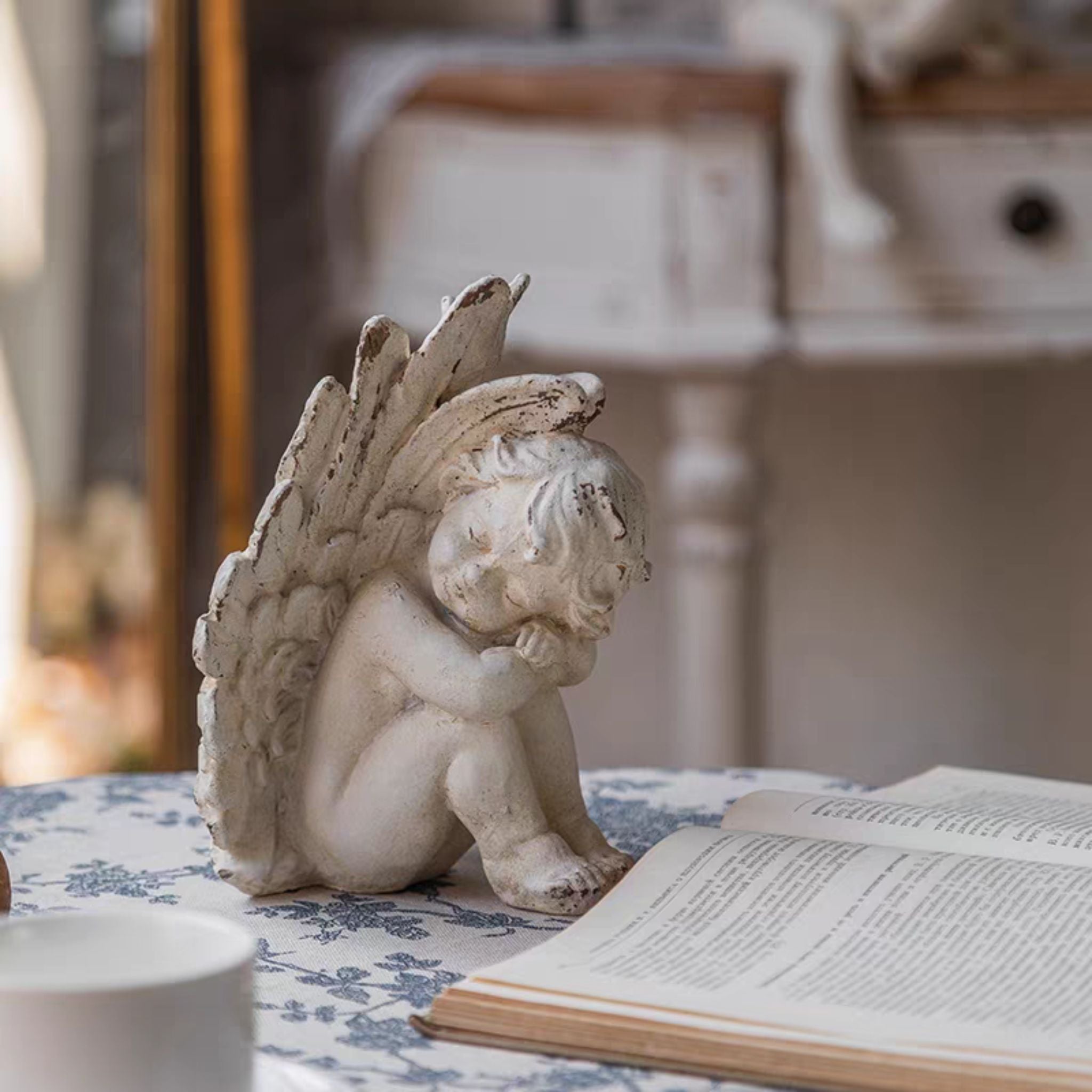 Romain French Provincial cherub figurine in distressed whitewash, curled with head bowed and wings fanned tall, styled on a blue toile tablecloth. Hero.
