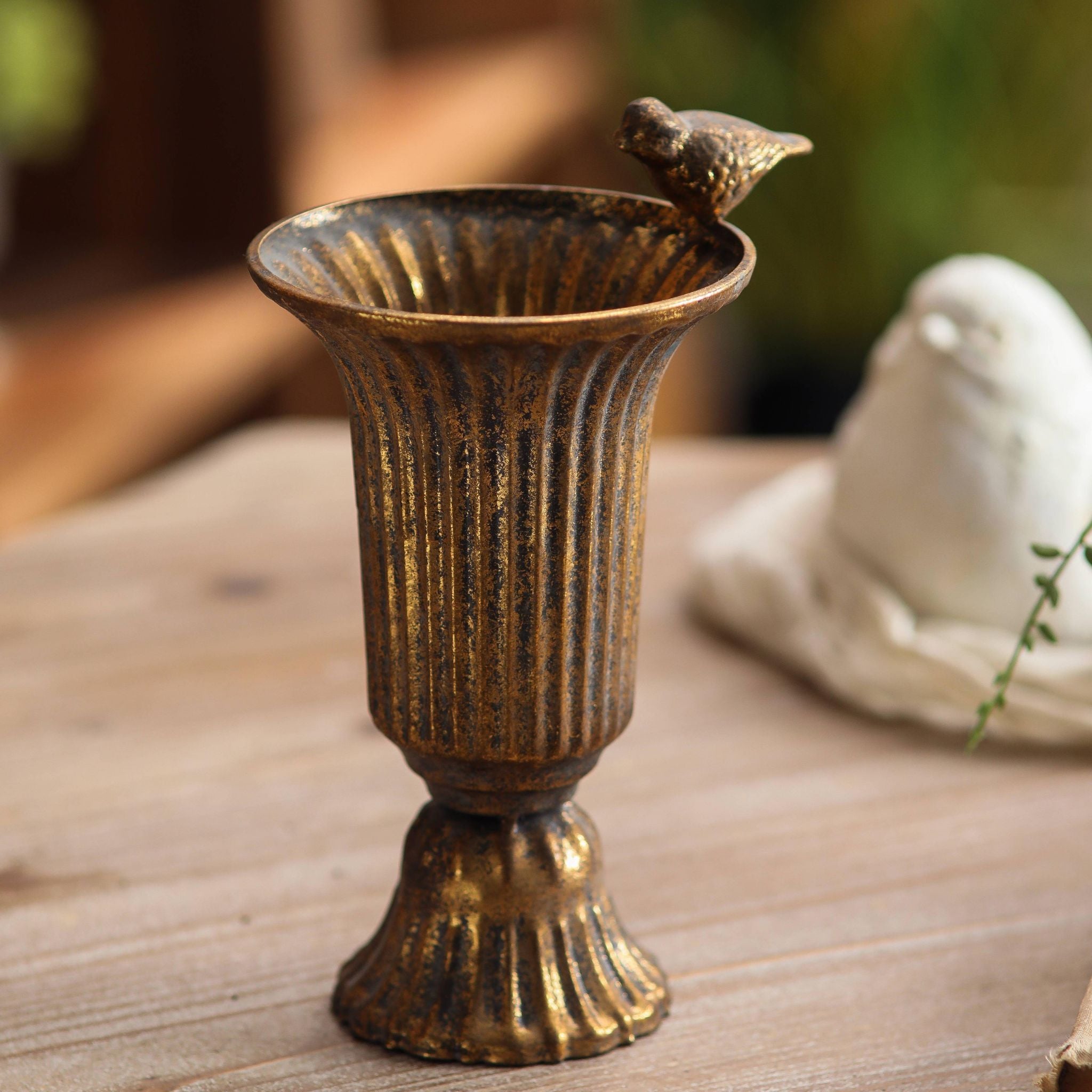 Valentine fluted urn vase in antique gold metal with a small cast bird on the rim, empty, on a timber table. Hero.