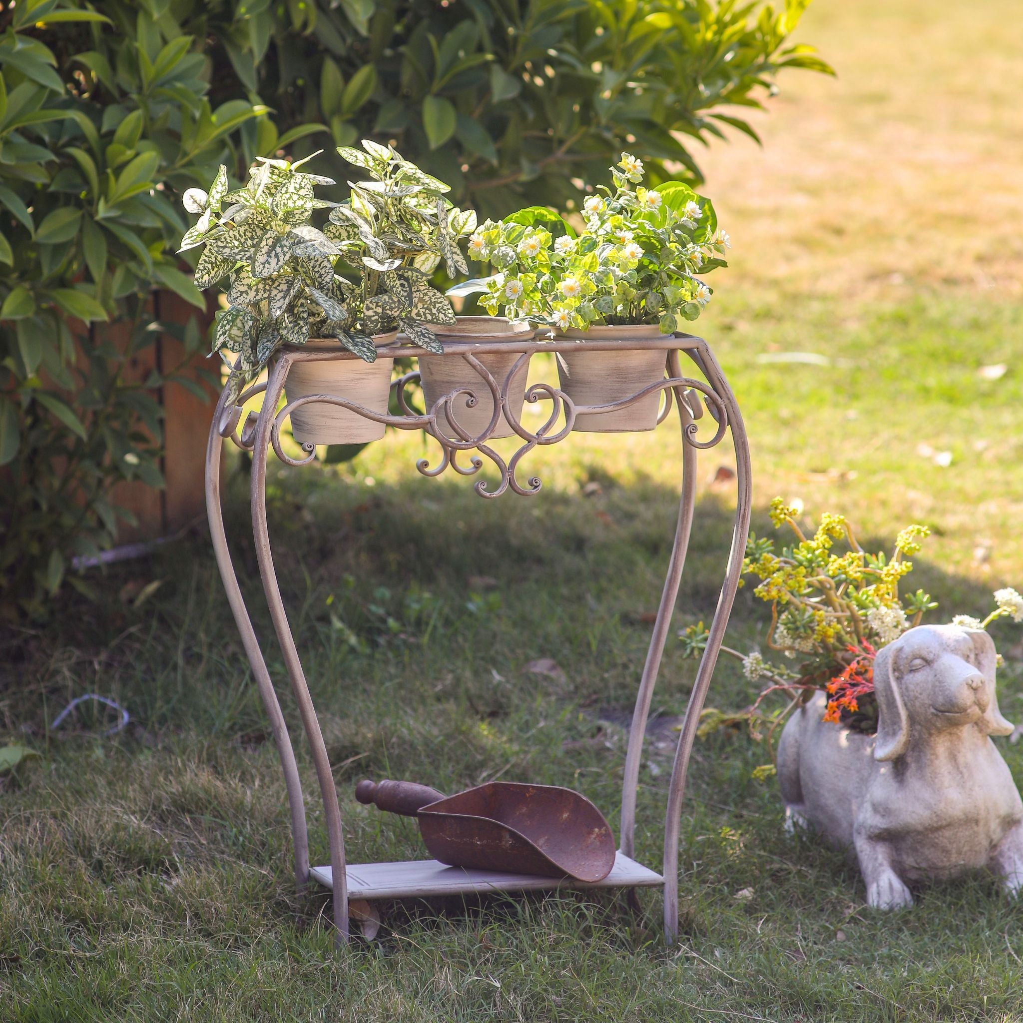 Yolande plant stand holding three potted plants, scrolled metal frame, set in a garden bed.